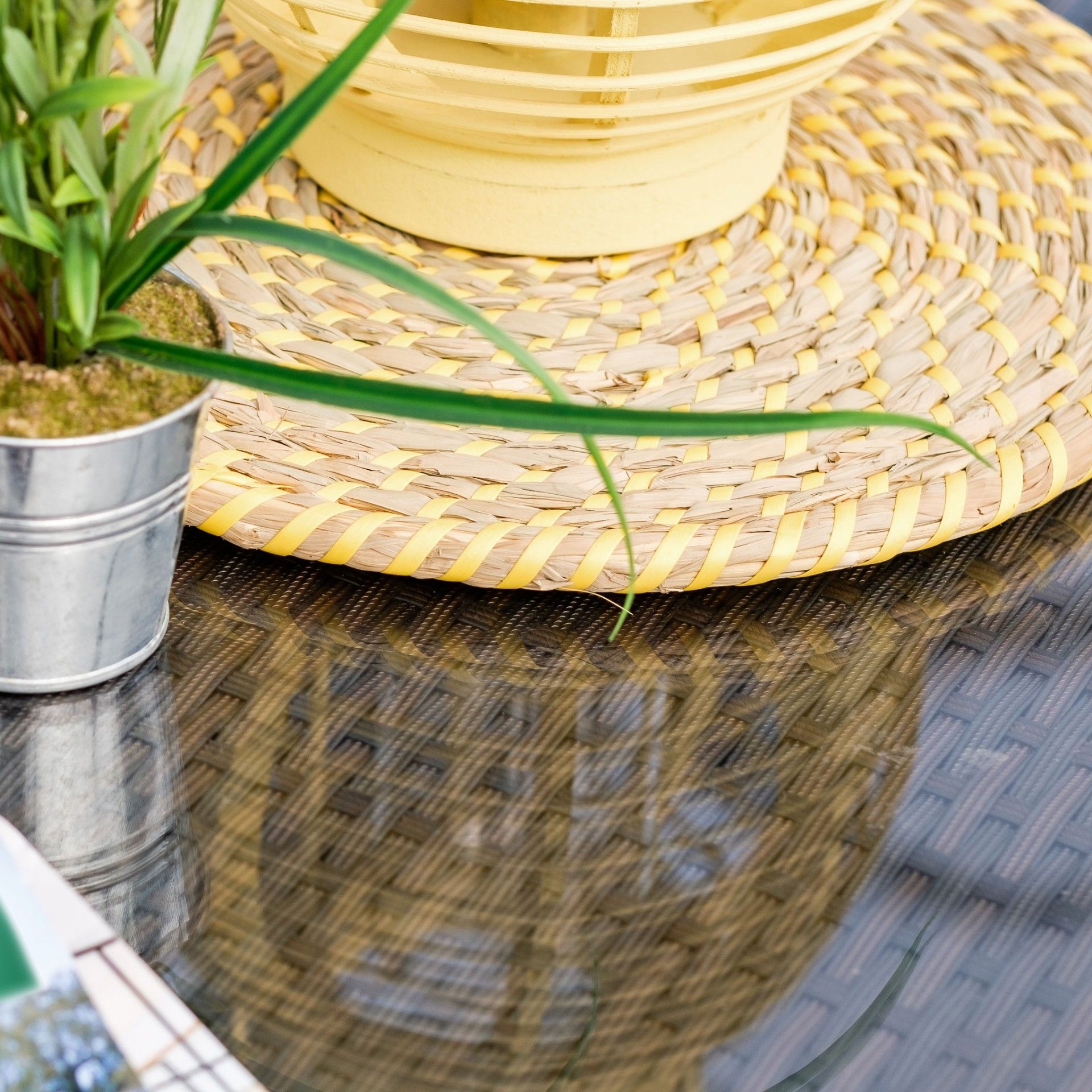 Mesa de vidro com cadeira de vime, tapete redondo tecido e vaso metálico com planta verde
