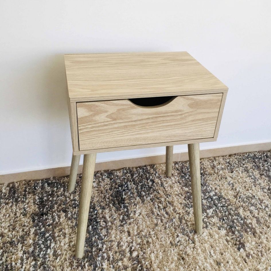 Mesa de cabeceira de madeira clara com gaveta e pernas finas sobre tapete castanho e cinza