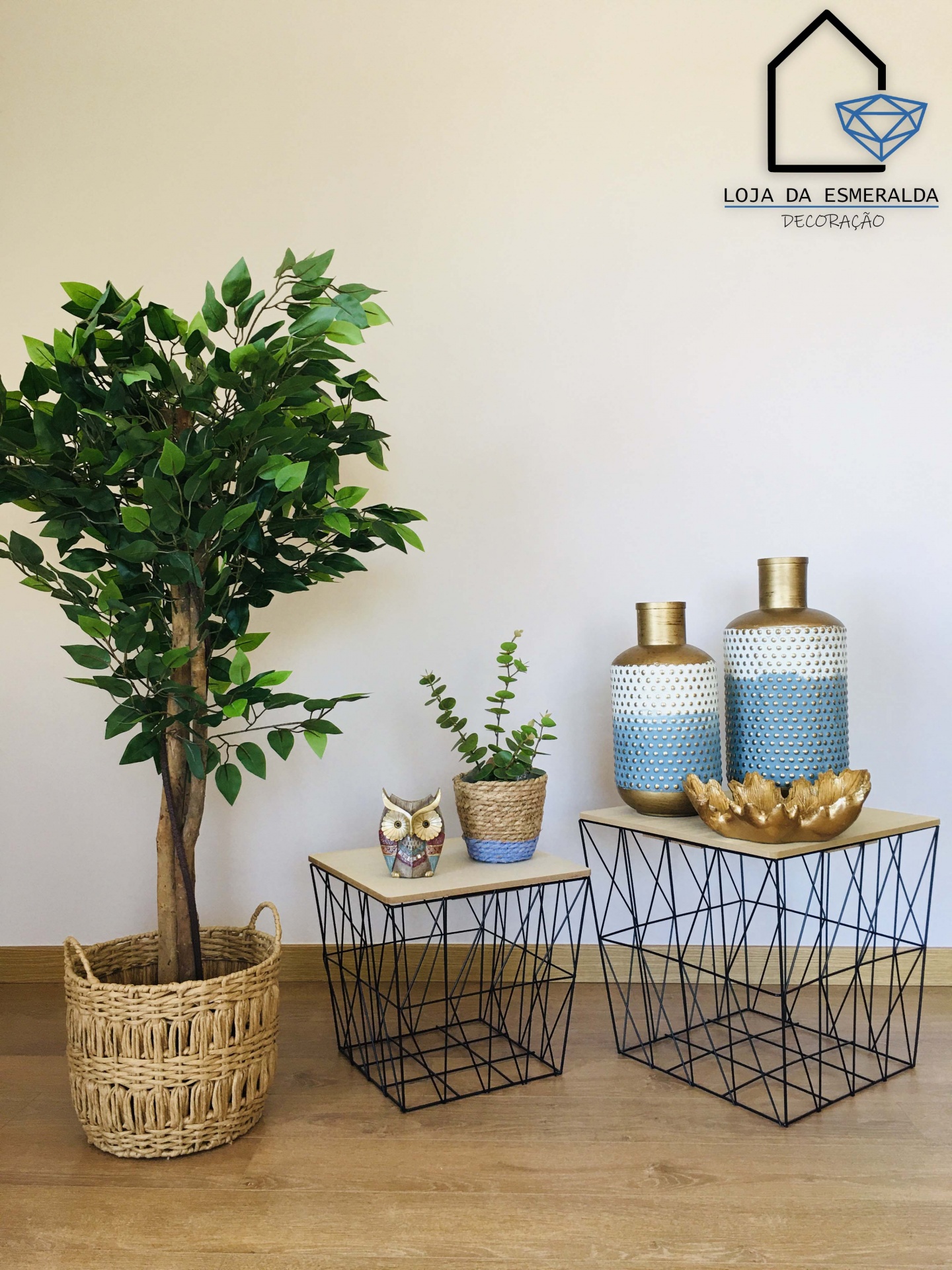Árvore artificial em vaso de vime ao lado de duas mesas com objetos decorativos e logo de loja na parede