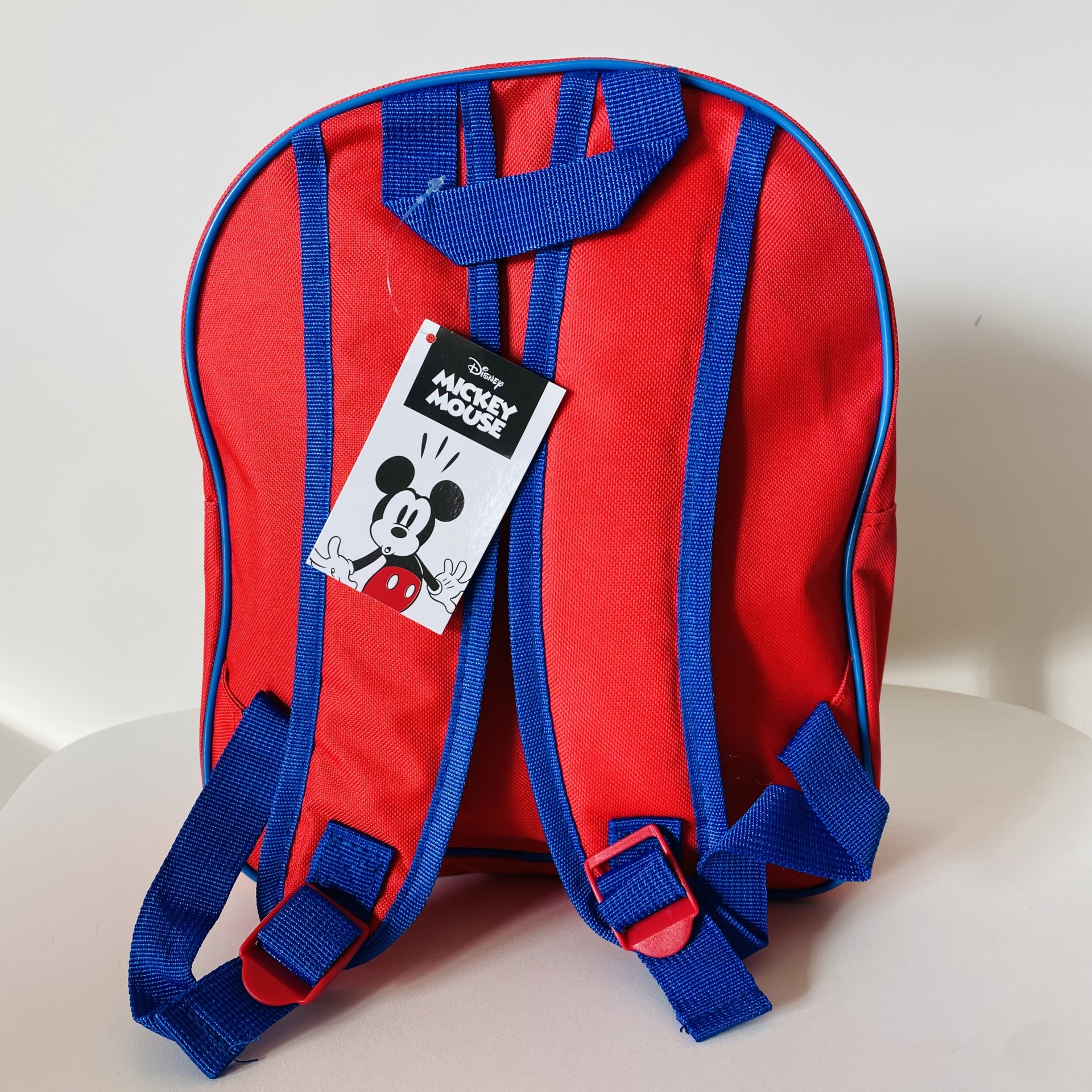 Mochila vermelha e azul com etiqueta do Mickey Mouse numa superfície branca