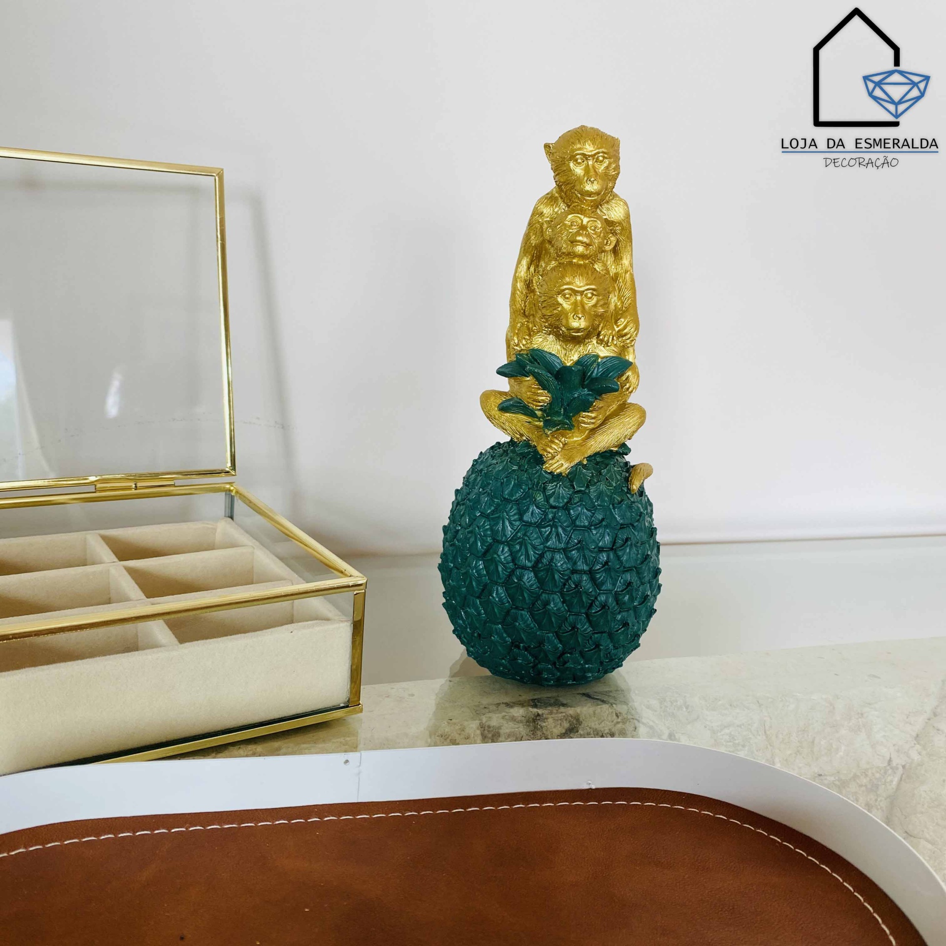 Estatueta com macacos dourados e base verde em forma de abacaxi sobre superfície com caixa transparente e bandeja