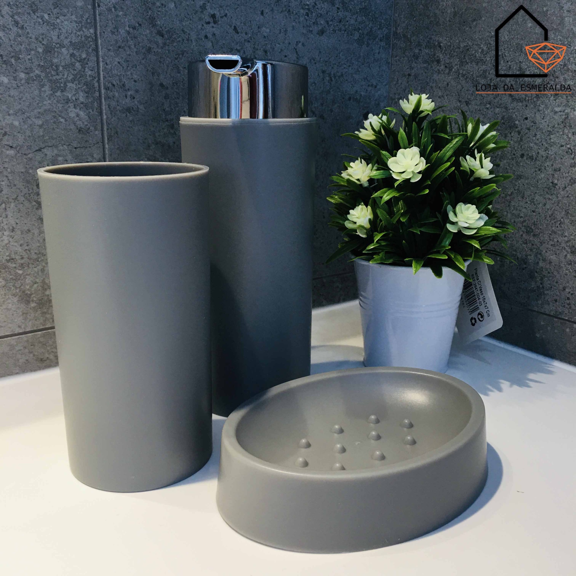 Conjunto de acessórios de casa de banho cinza com dispensador, copo e saboneteira ao lado de vaso com flores artificiais.