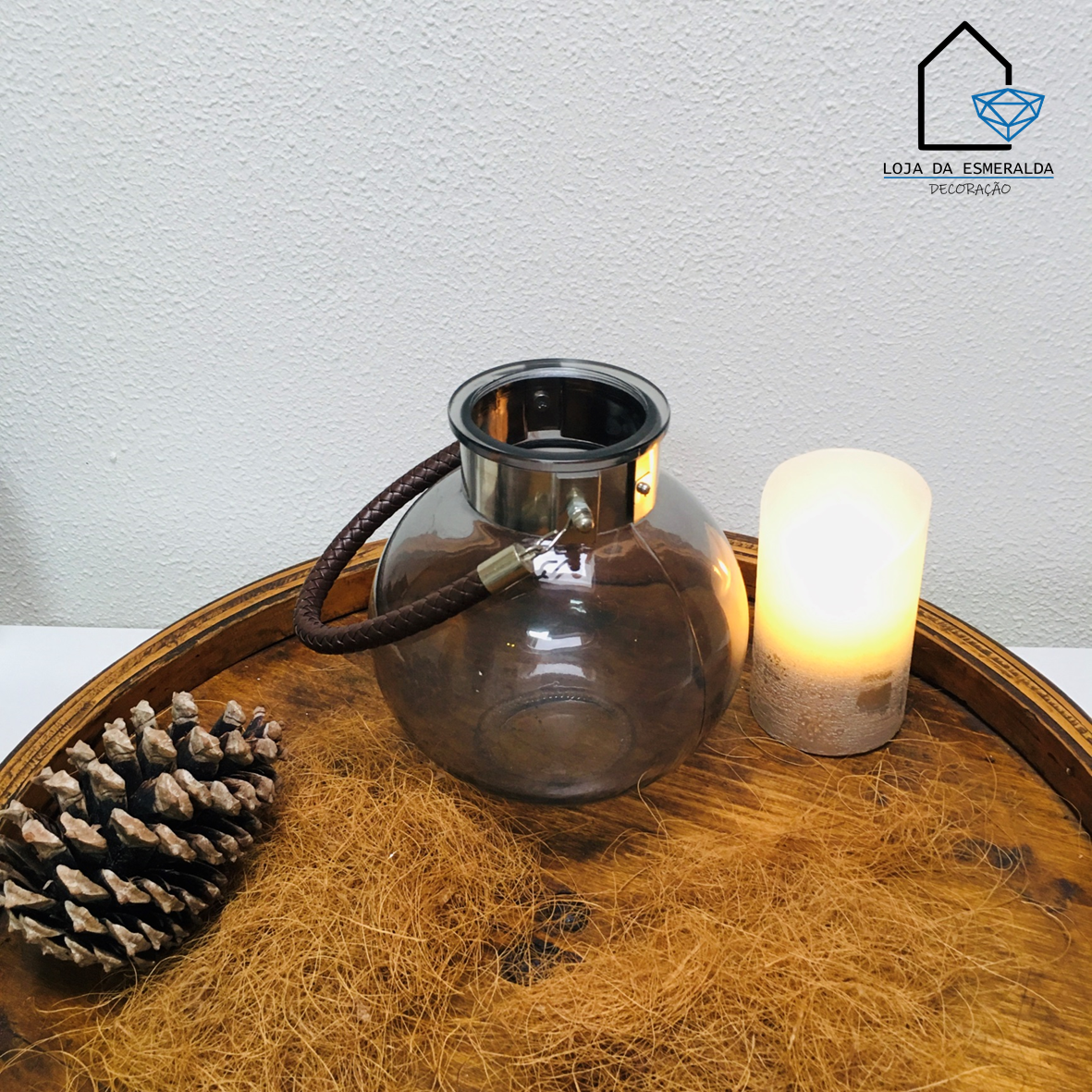 Lanterna de vidro com aro metálico e alça castanha junto a vela acesa e pinheiro seco numa bandeja de madeira