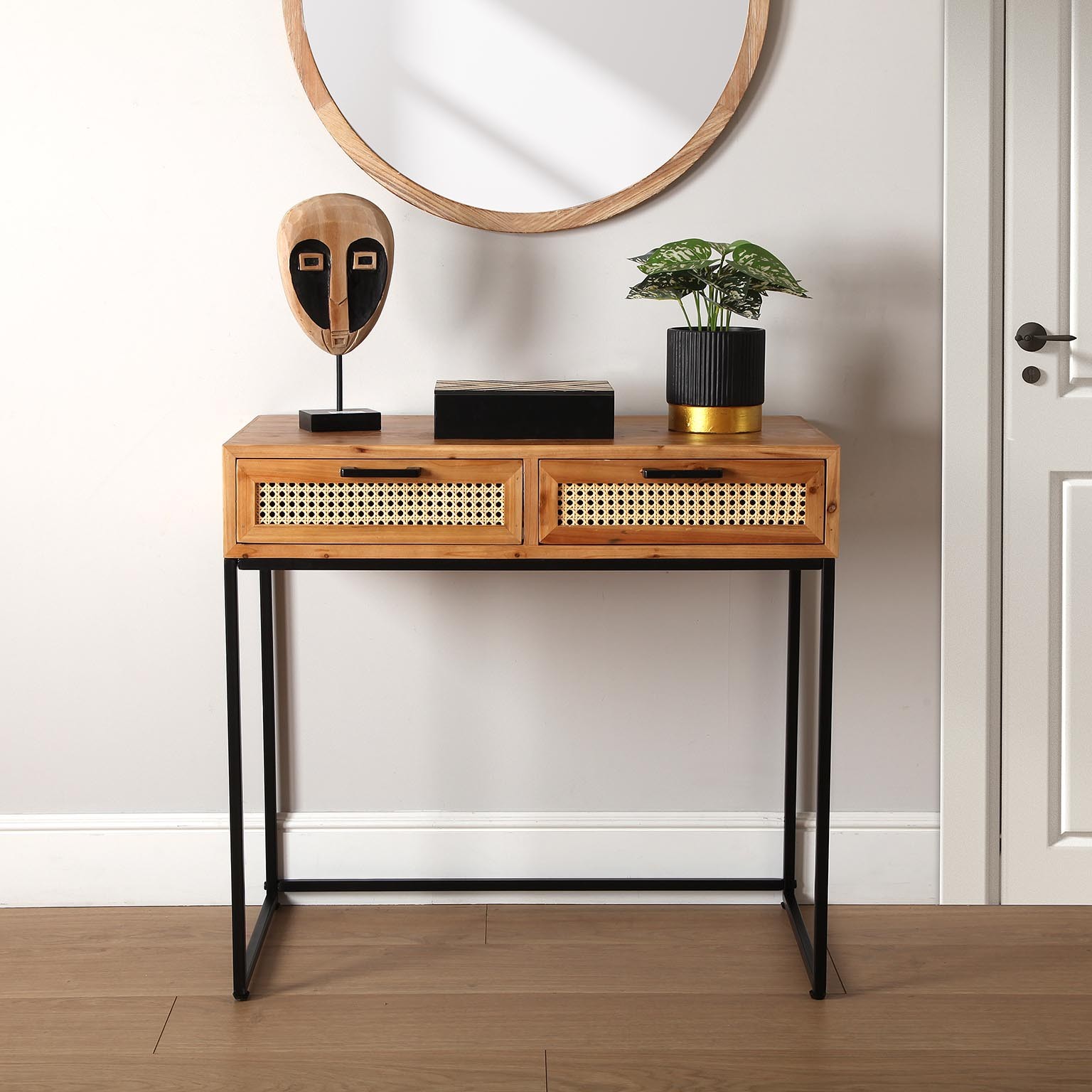 Aparador de madeira clara com duas gavetas de palhinha e estrutura preta, decorado com máscara, caixa preta e planta, contra parede branca e porta ao lado