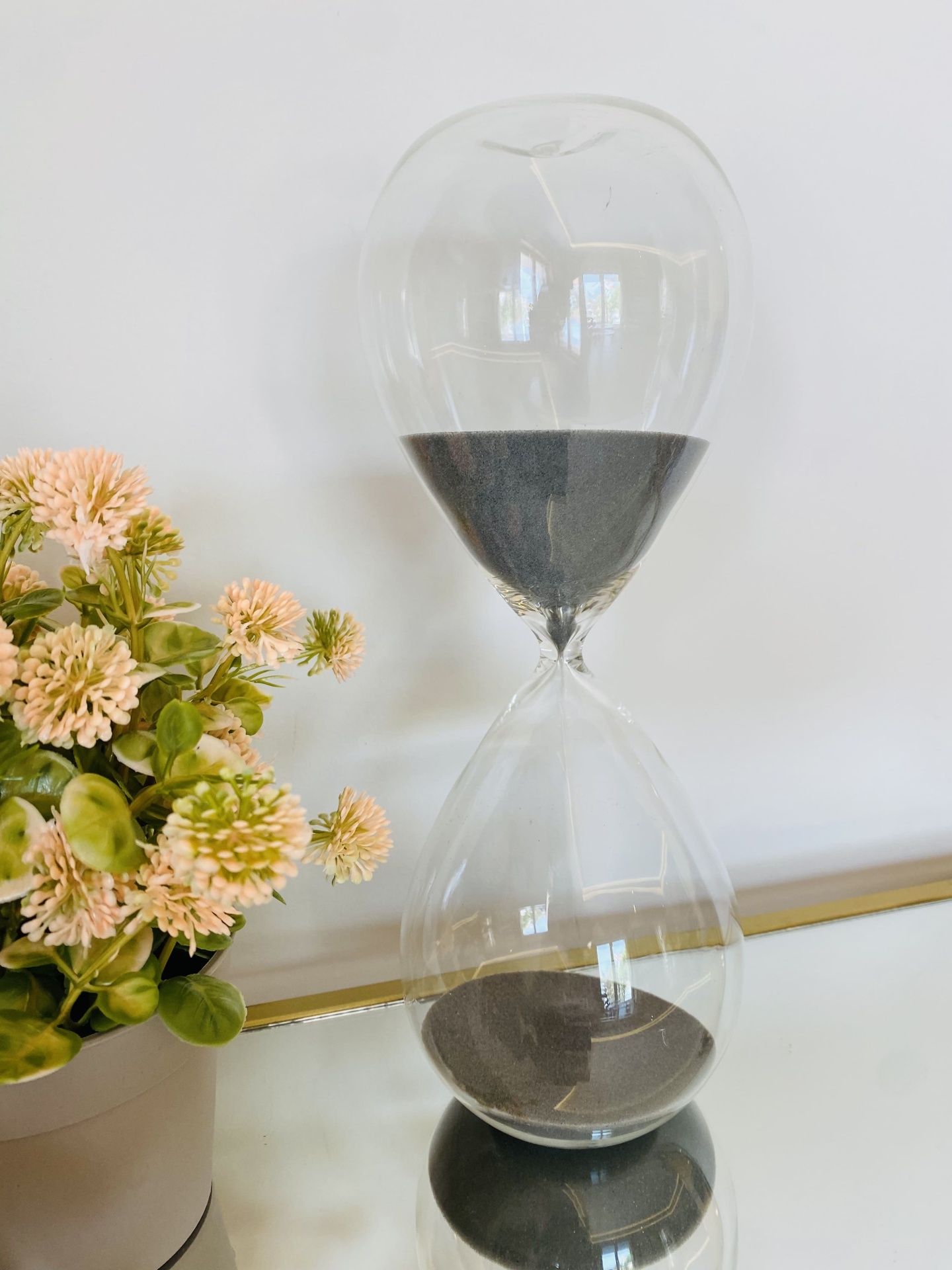 Relógio de areia transparente com areia preta ao lado de planta com flores cor de rosa claro