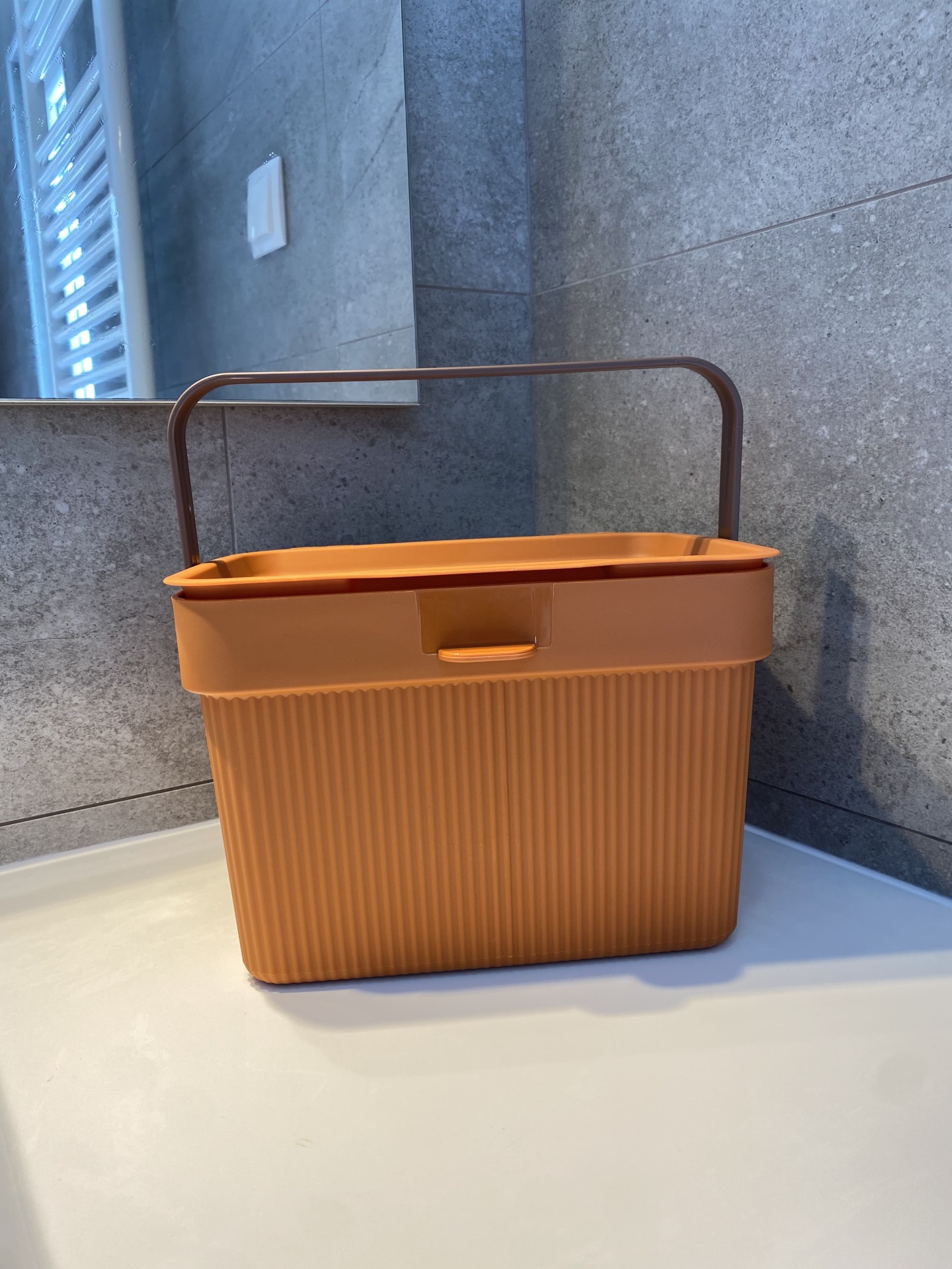 Cesto de plástico castanho com alça numa bancada branca e paredes cinzentas