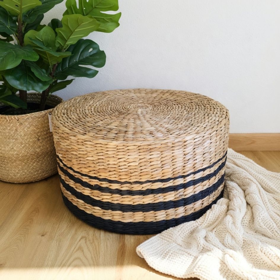Pufe redondo em palha natural com faixas pretas e manta creme ao lado.