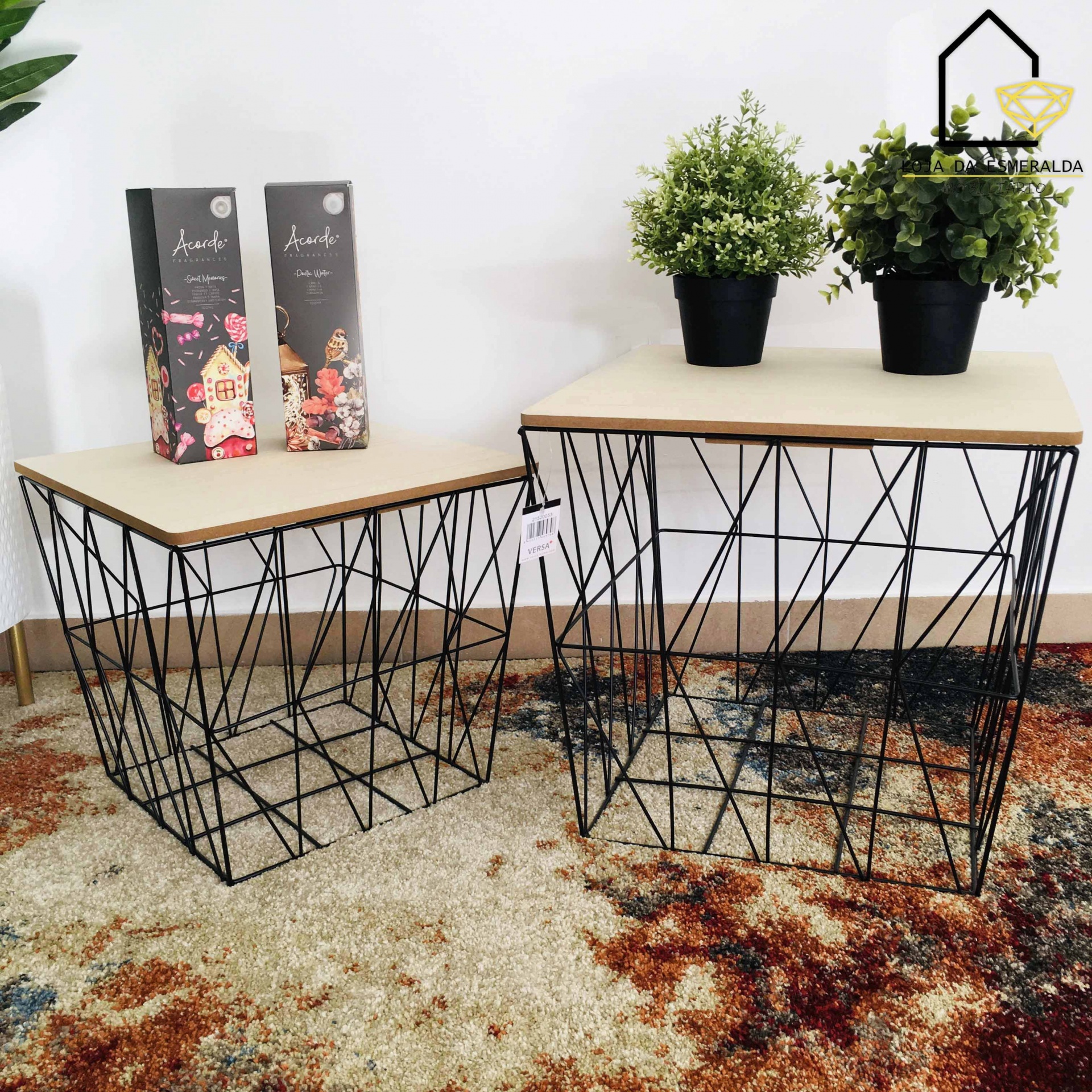 Mesas de centro com estrutura metálica preta geométrica e tampo de madeira clara, sobre tapete colorido, com plantas em vasos pretos e embalagens decorativas ao fundo