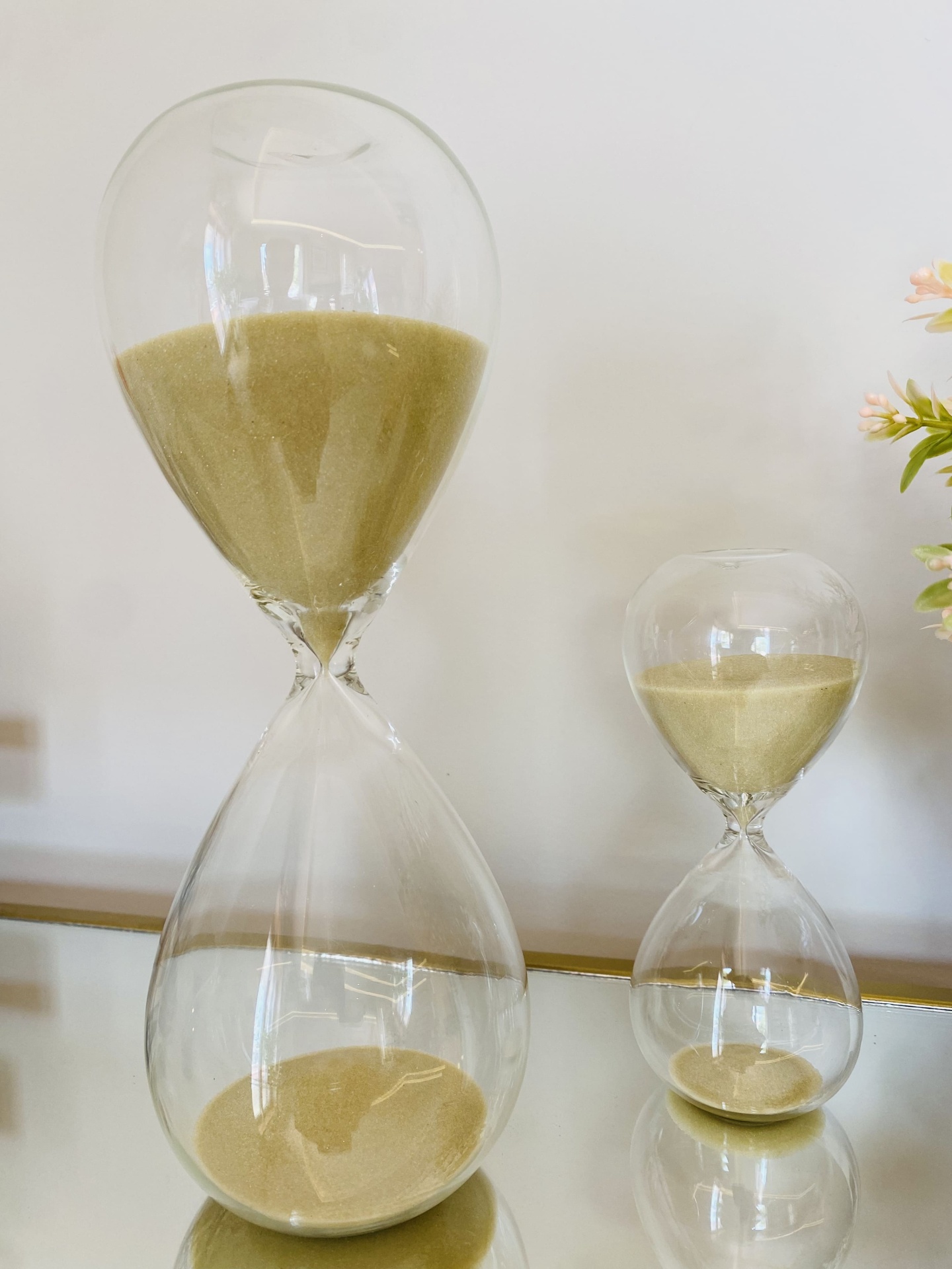 Dois relógios de areia transparentes, um grande e outro pequeno, com areia amarela clara sobre superfície espelhada