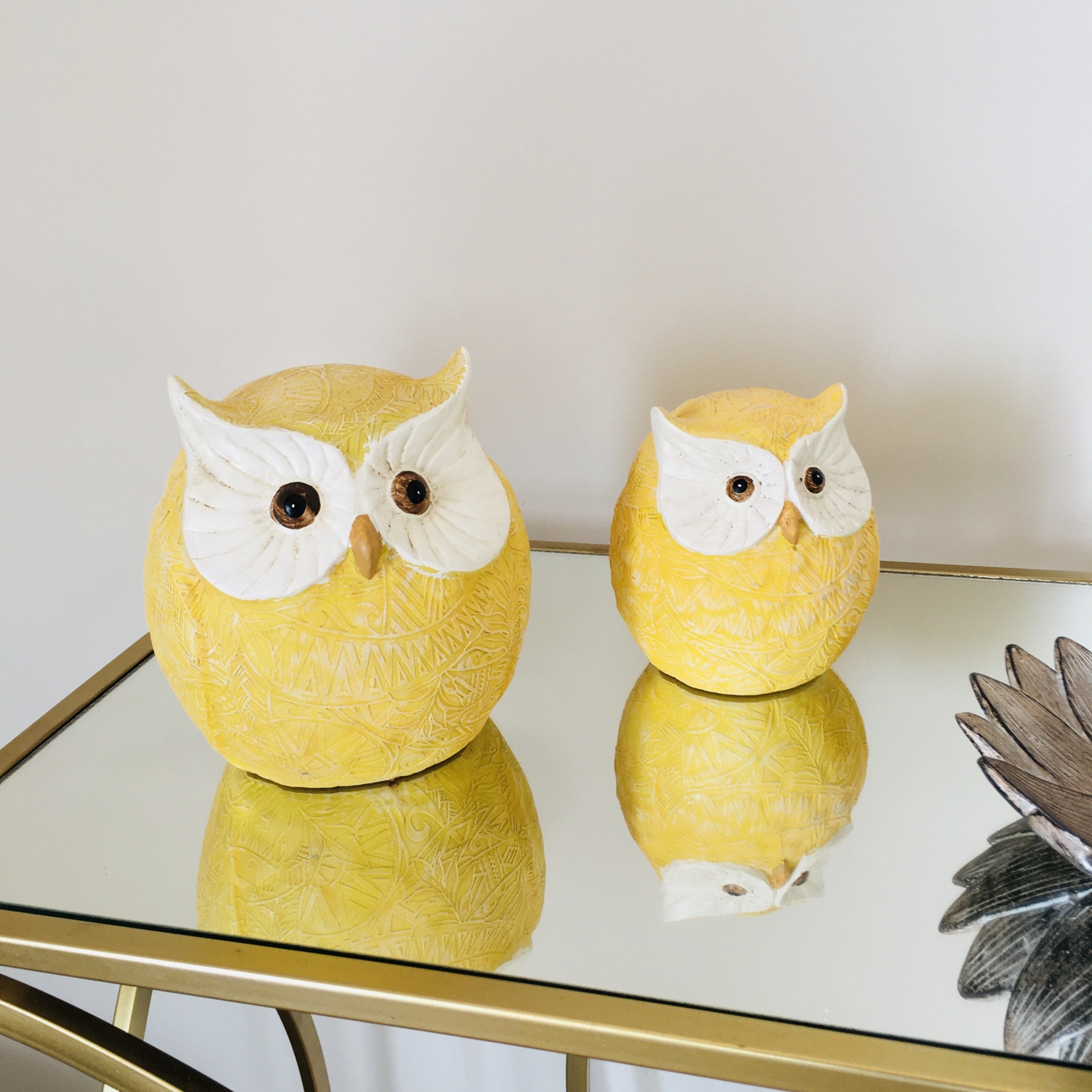 Duas corujas decorativas amarelas com olhos brancos sobre mesa de vidro espelhado com estrutura dourada