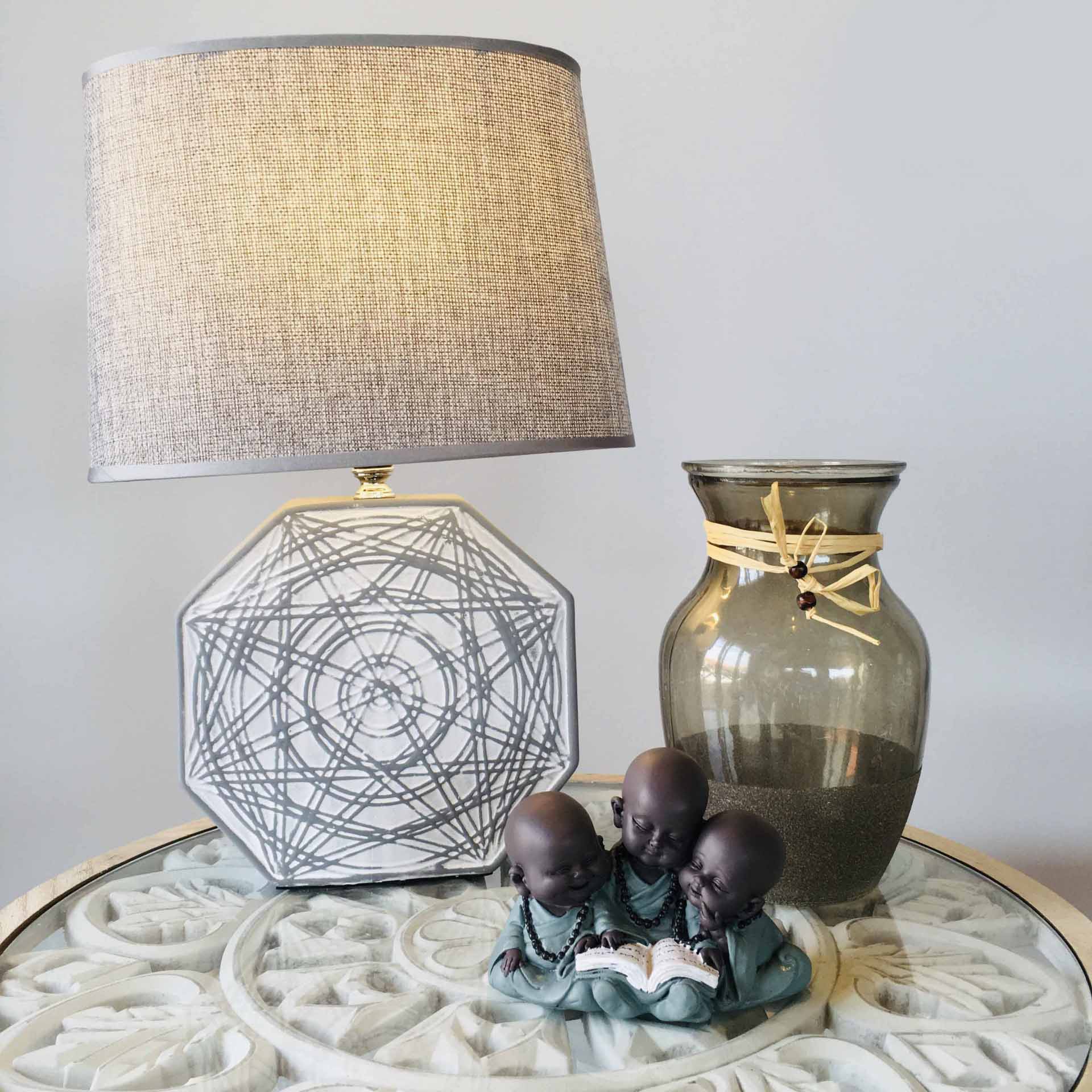 Lâmpada de mesa, vaso de vidro e estatueta de crianças lendo, sobre mesa branca com padrão floral.