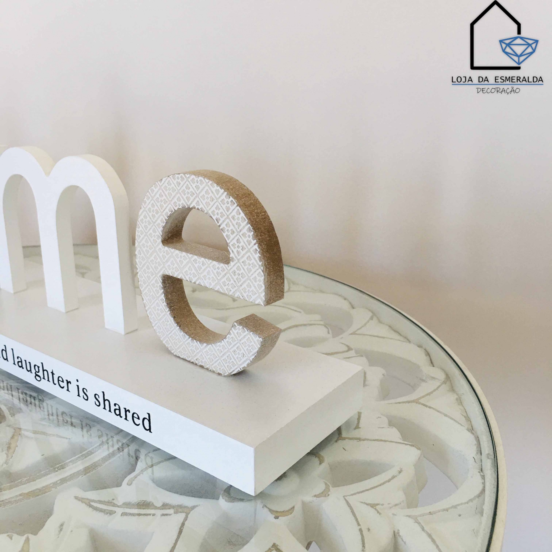 Objeto decorativo em madeira com letras brancas e textura geométrica sobre mesa de vidro com detalhes florais
