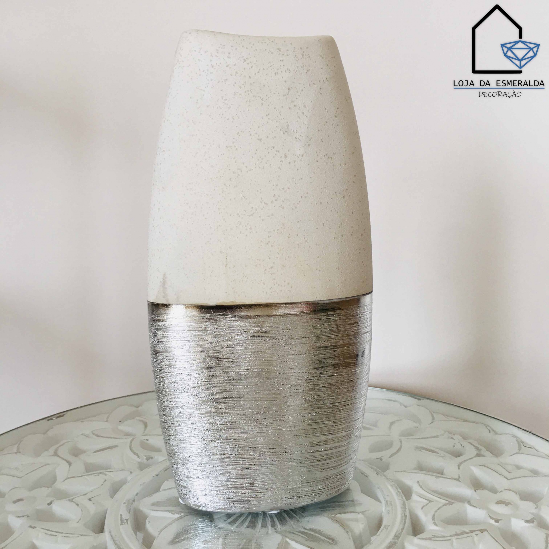 Vaso decorativo branco e prateado sobre mesa de vidro com padrão floral e logotipo da loja ao fundo