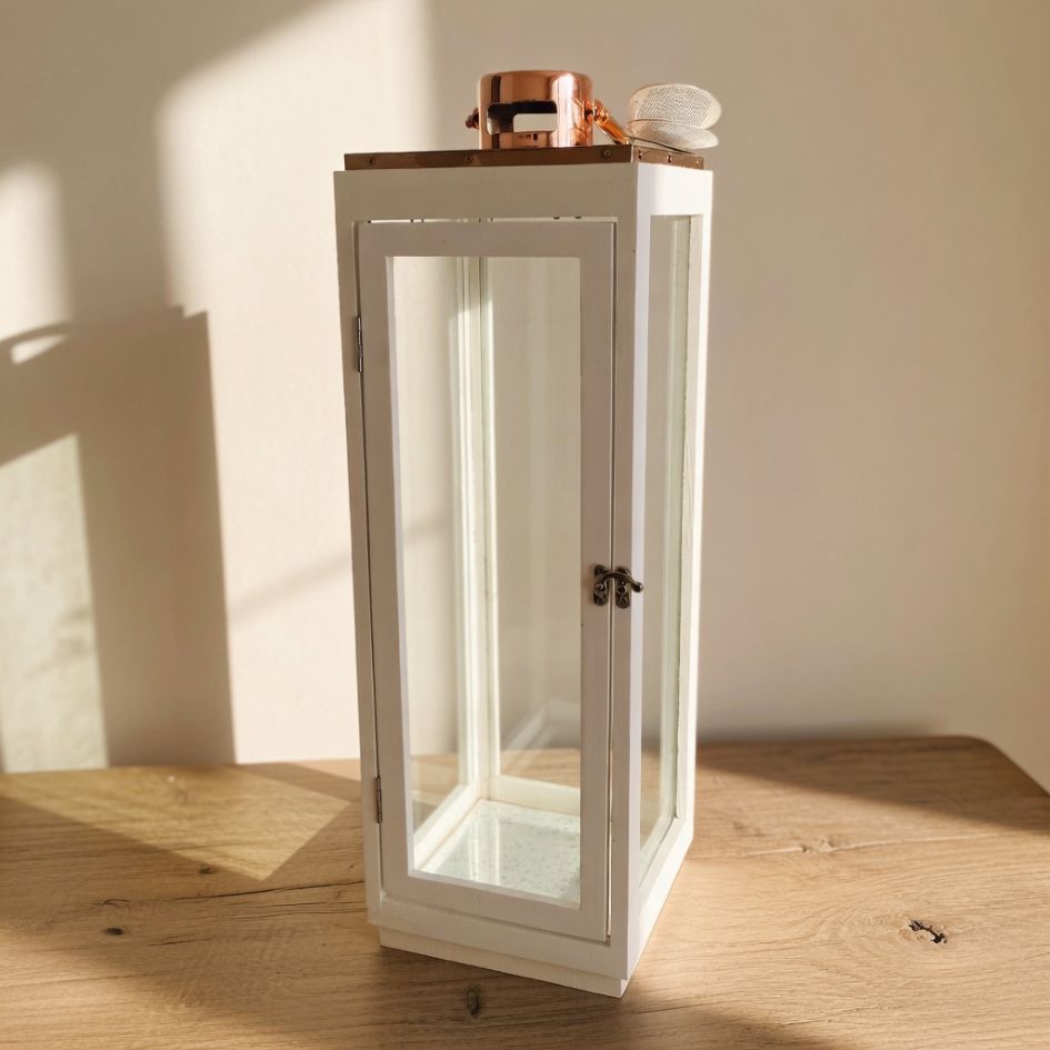 Lanterna decorativa branca em madeira e vidro com topo em cobre sobre mesa de madeira