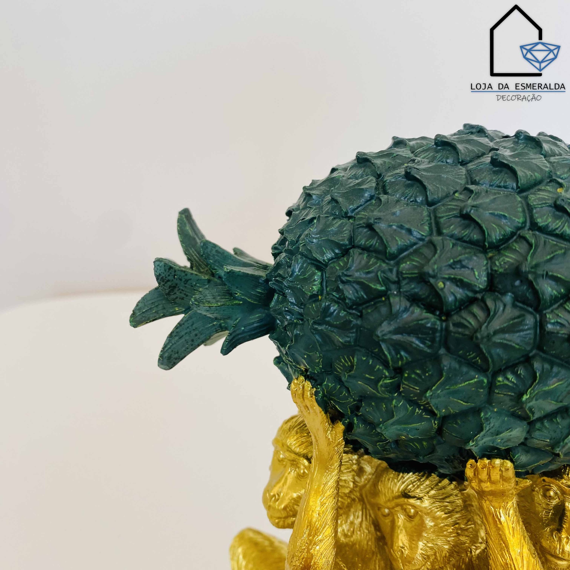 Escultura de quatro macacos dourados a segurar um abacaxi verde escuro, fundo branco com logotipo.