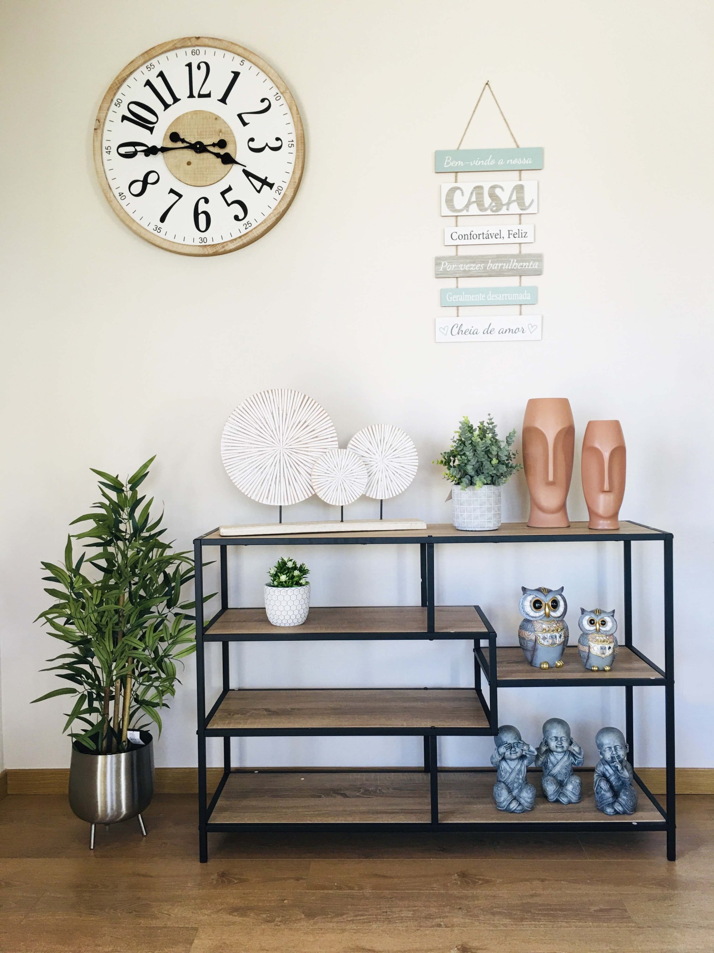ambiente interior com relógio de parede, placa decorativa, estante com plantas e esculturas diversas