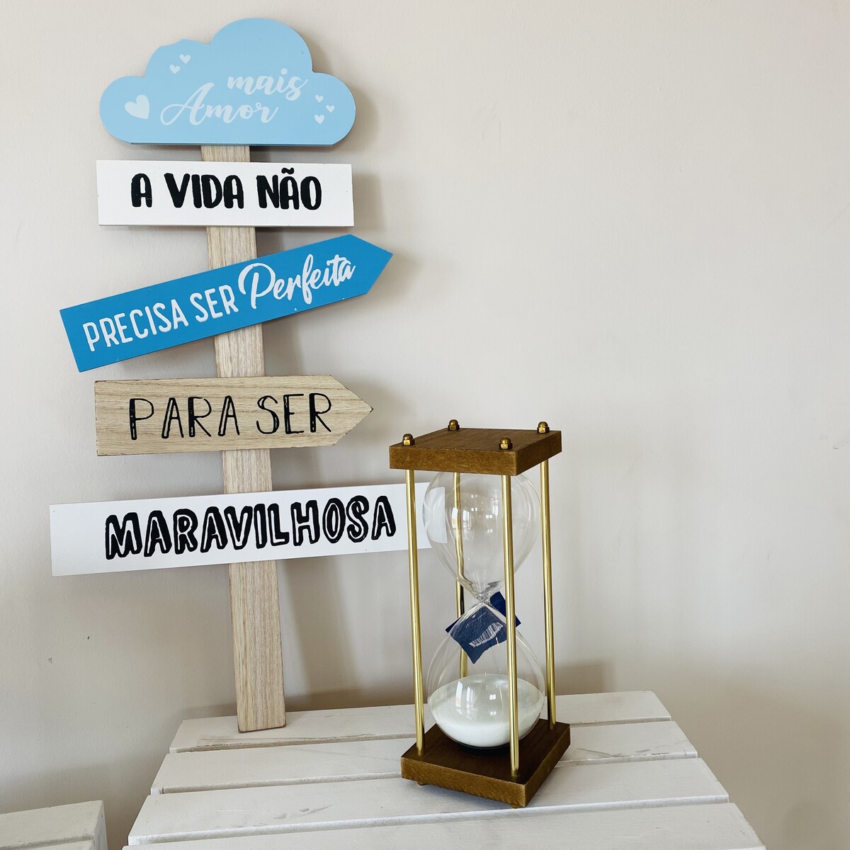 Relógio de areia com base dourada junto a placa decorativa de madeira com frases em português