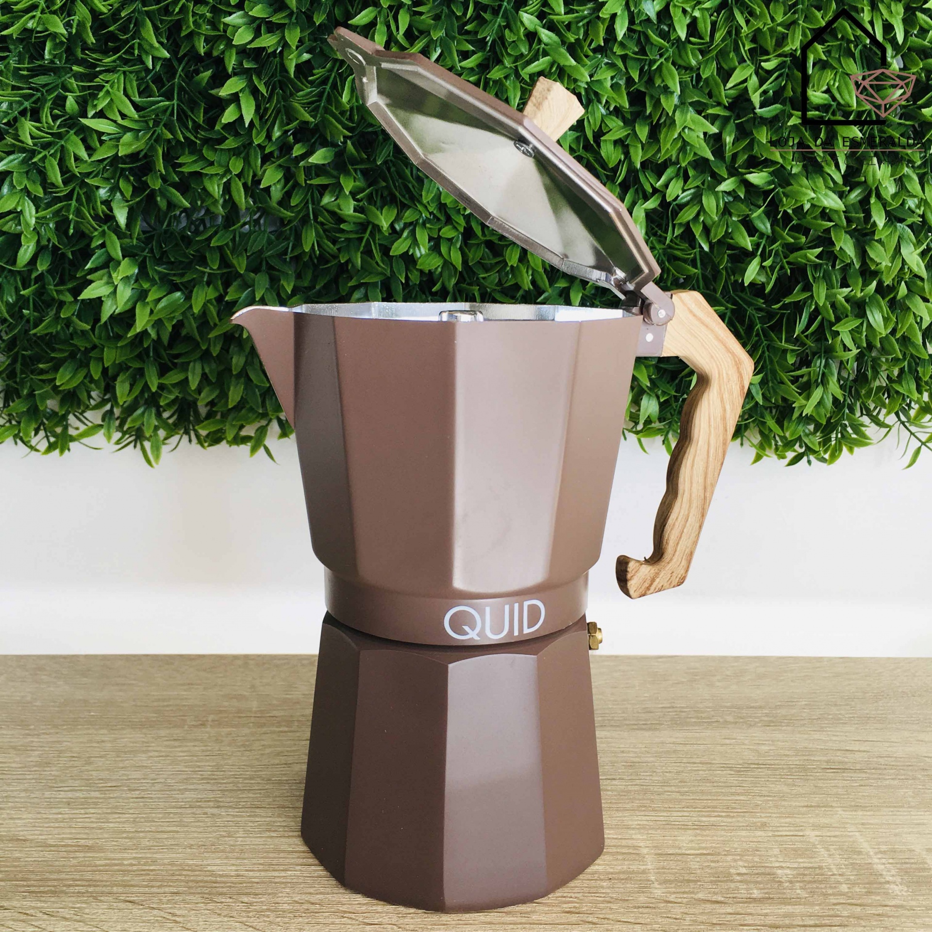 Cafeteira italiana castanha com pega de madeira sobre mesa clara e fundo de folhas verdes