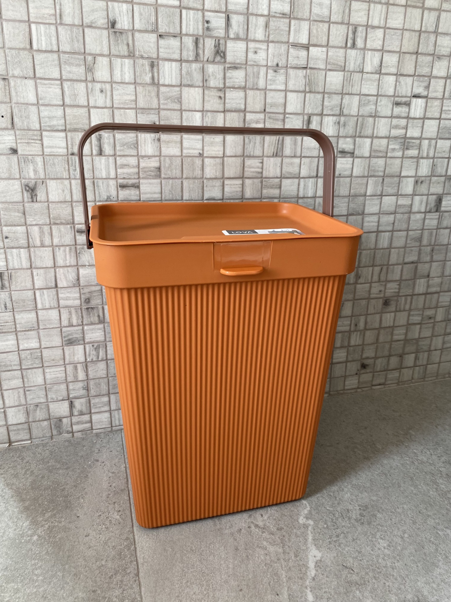 Contentor de plástico laranja com pega castanha sobre chão e parede de azulejos cinzentos