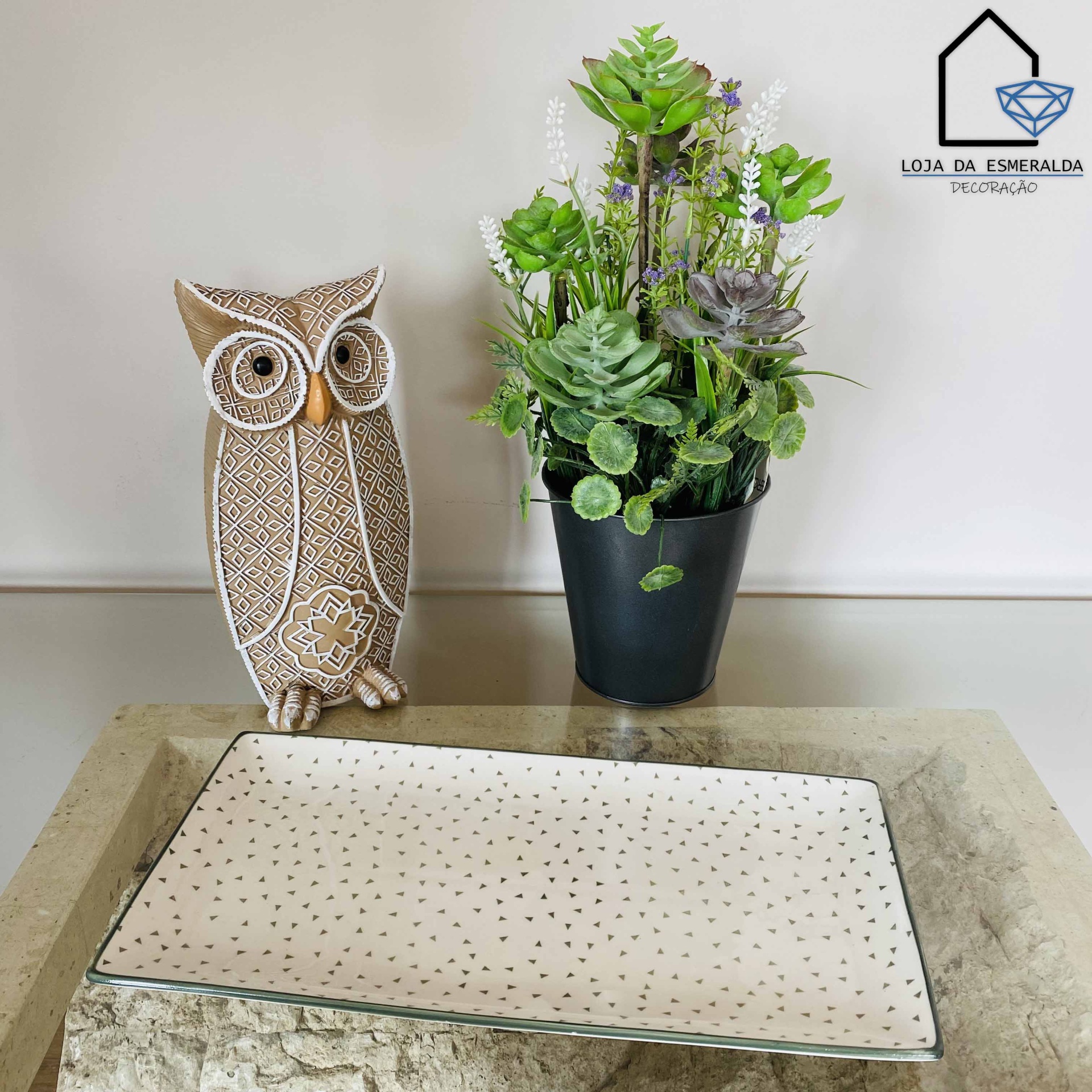 Decoração com coruja de padrão geométrico, vaso preto com plantas artificiais e tabuleiro branco com padrões pequenos.