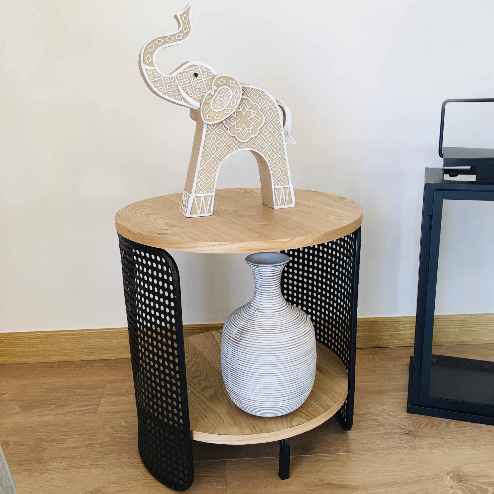Mesa redonda de apoio com tampo e prateleira em madeira clara e estrutura metálica preta, com elefante decorativo e vaso cerâmico
