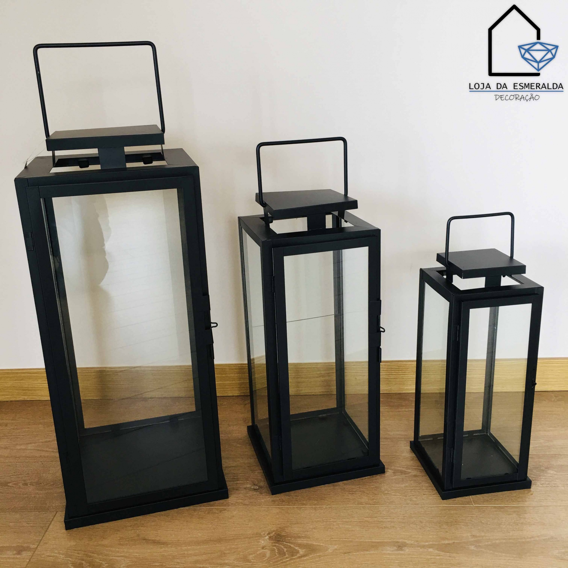 Três candeeiros de metal pretos com vidro em diferentes tamanhos em chão de madeira clara