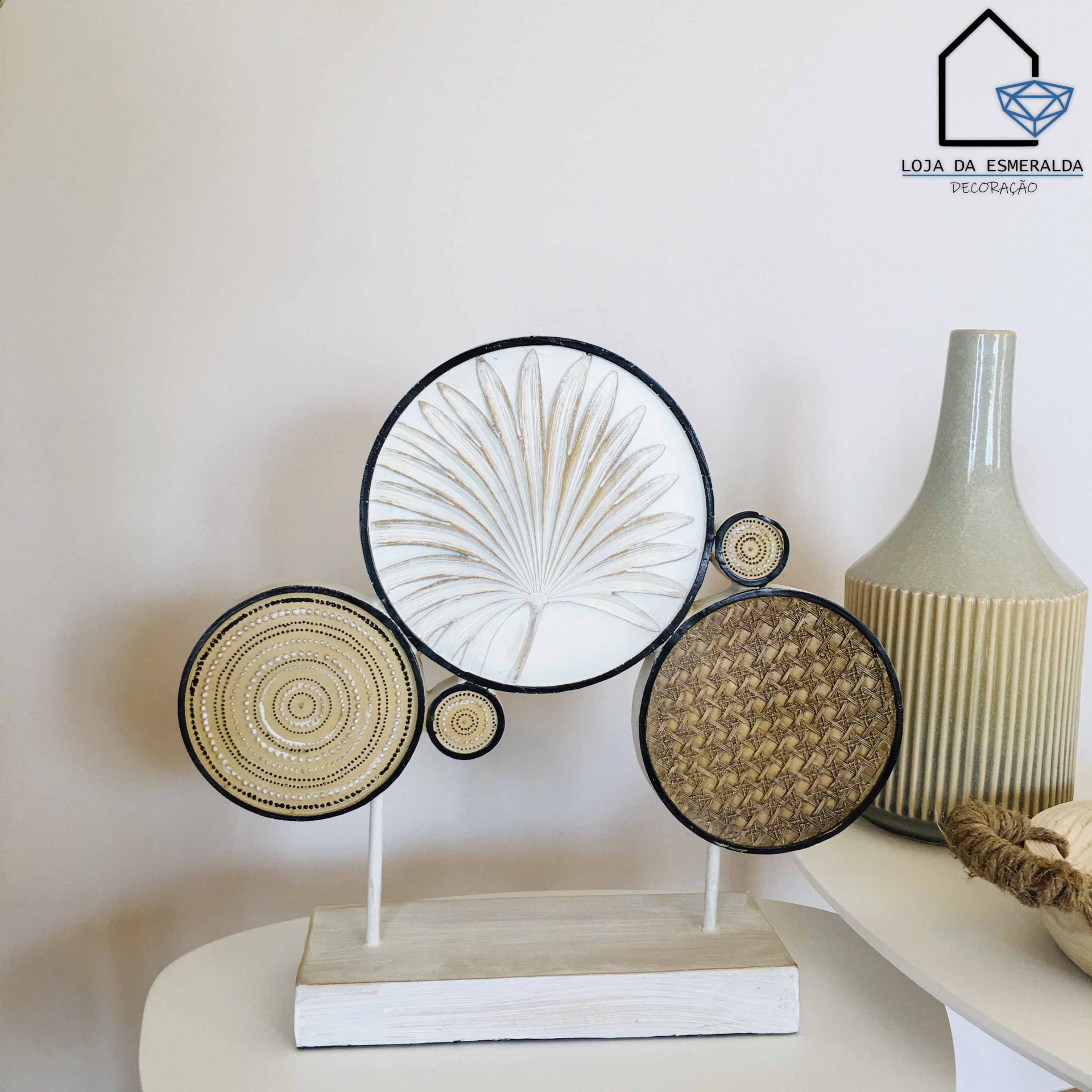 Escultura decorativa de metal com círculos texturizados e uma folha branca, sobre base branca, ao lado de vaso bege em superfície branca