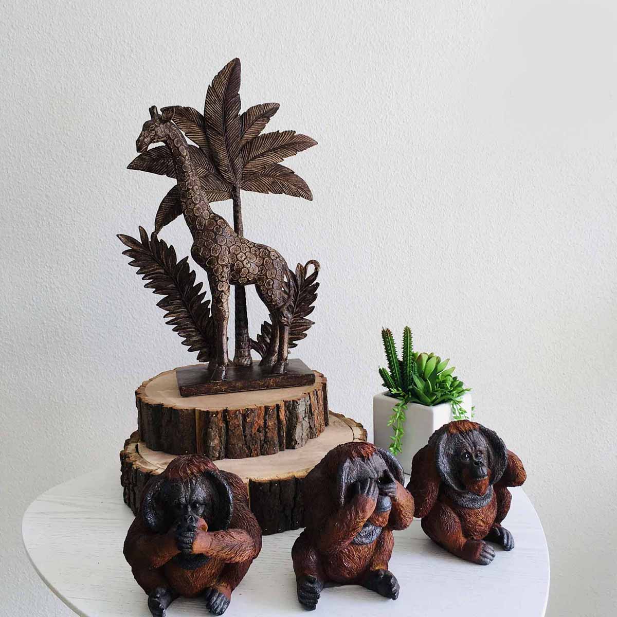 Três estatuetas de macacos em madeira com pose dos três macacos sábios e uma escultura de girafa em madeira com folhas, sobre uma mesa branca