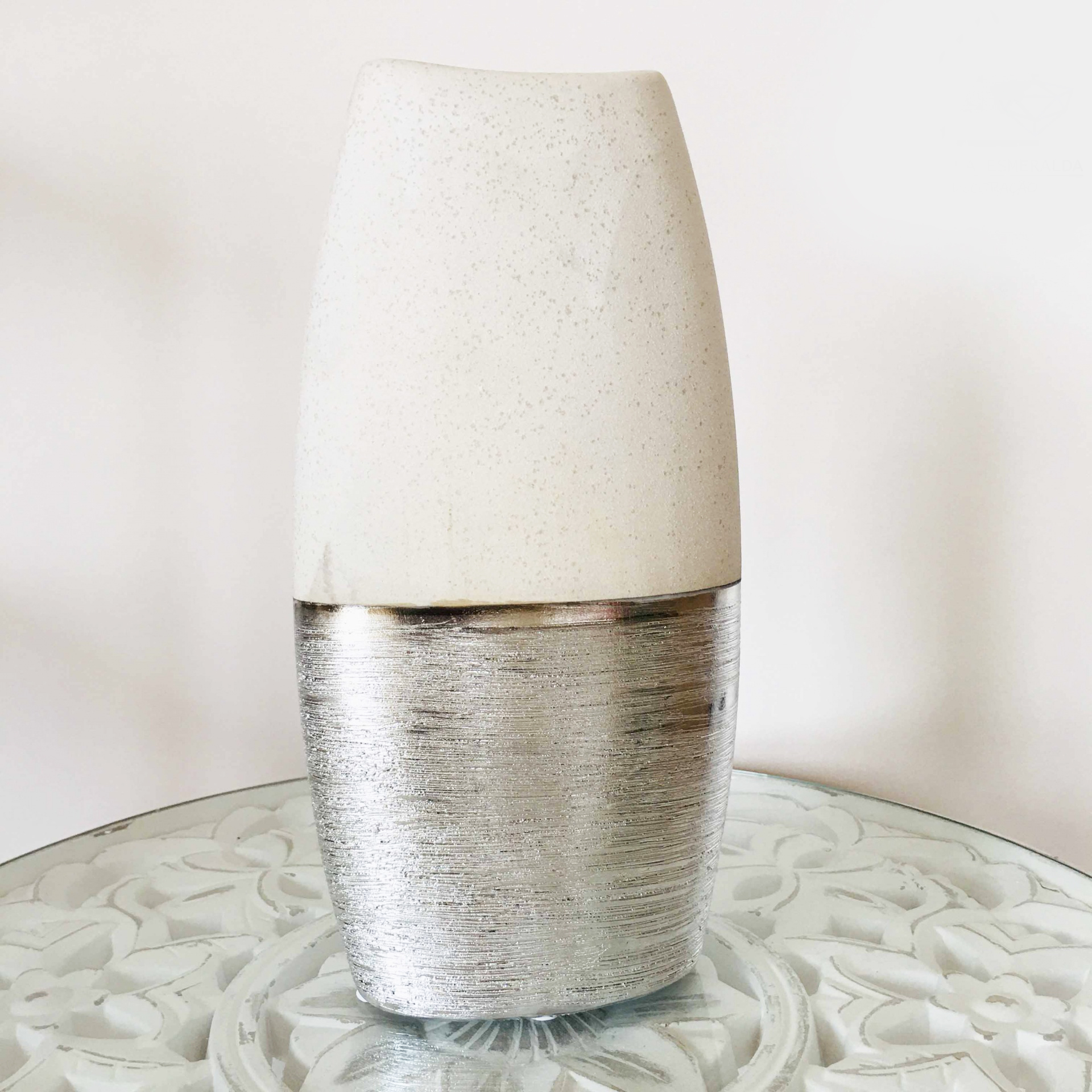 Vaso decorativo em prateado e branco sobre mesa de vidro com padrão floral