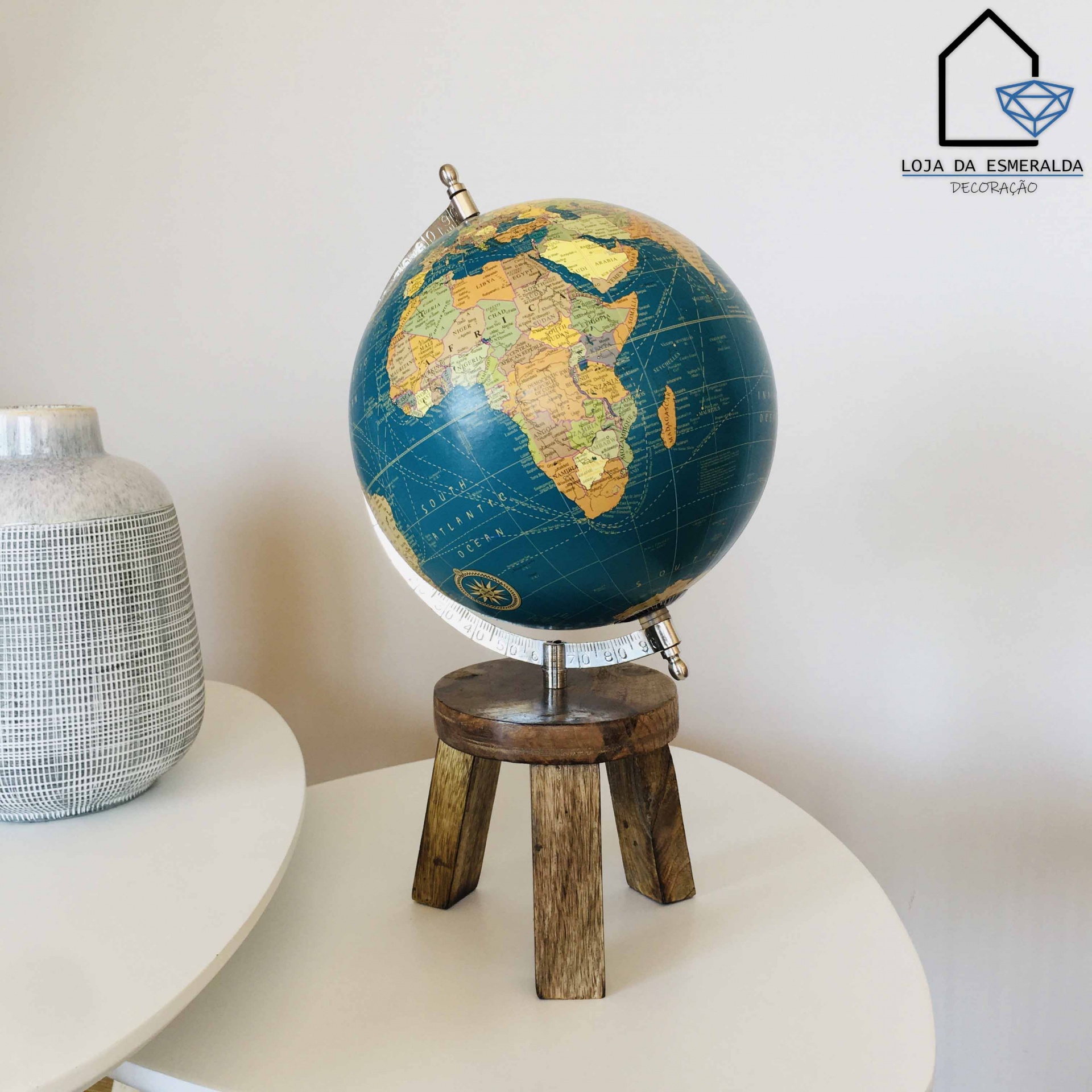 Globo terrestre decorativo com base de madeira numa mesa branca ao lado de um vaso. 