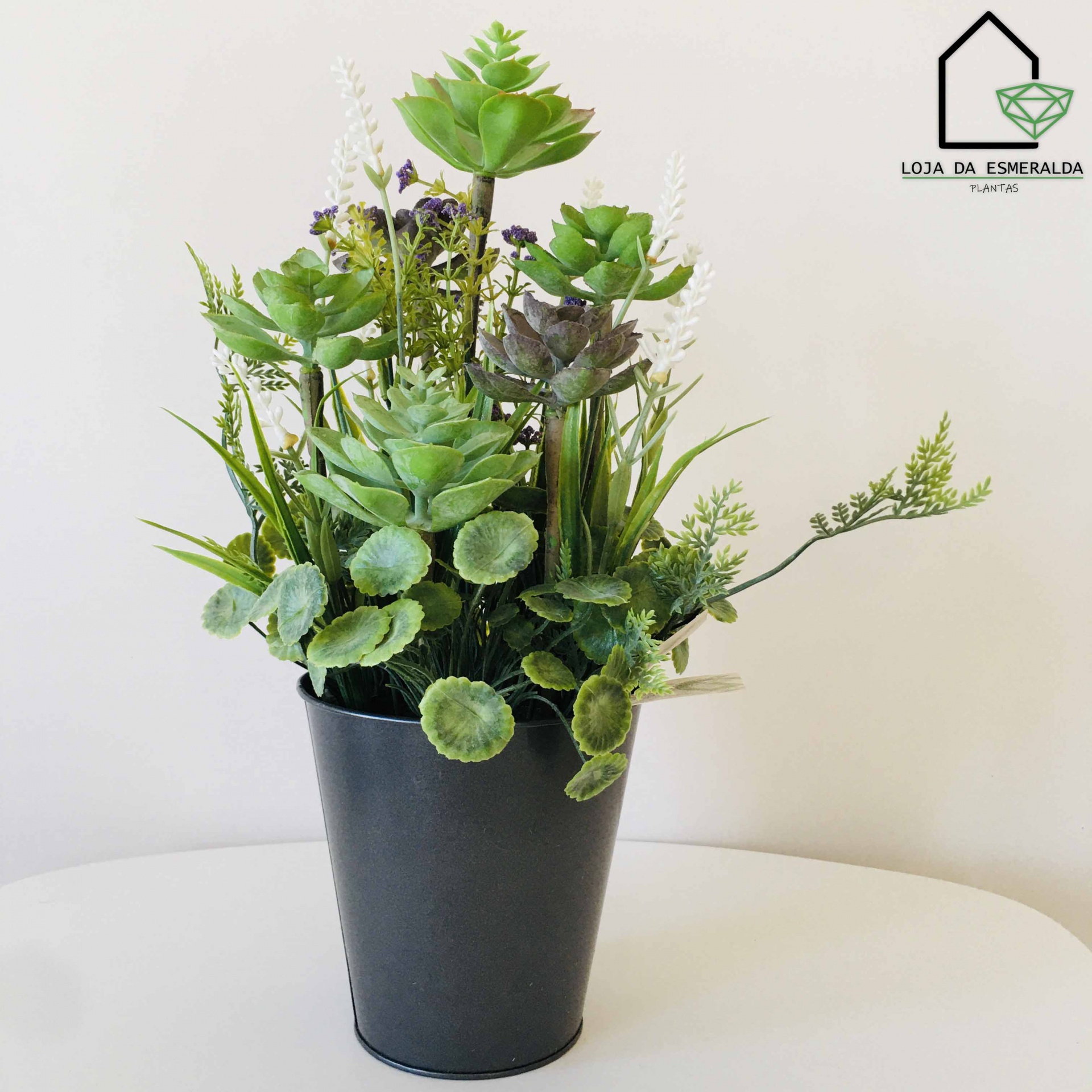 Arranjo de plantas artificiais em vaso preto sobre superfície branca.