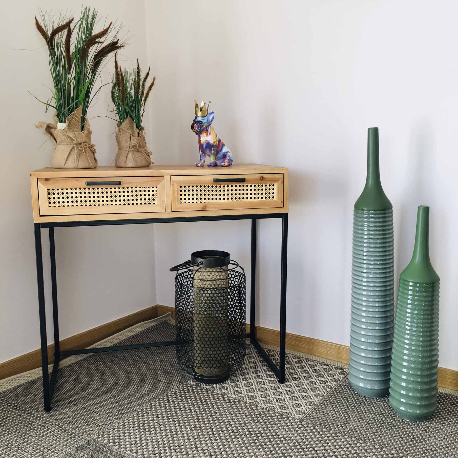 Mesa com gavetas em madeira e metal, vasos com plantas e estatueta colorida em cima, candeeiro e jarras verdes grandes ao lado, sobre carpete cinza
