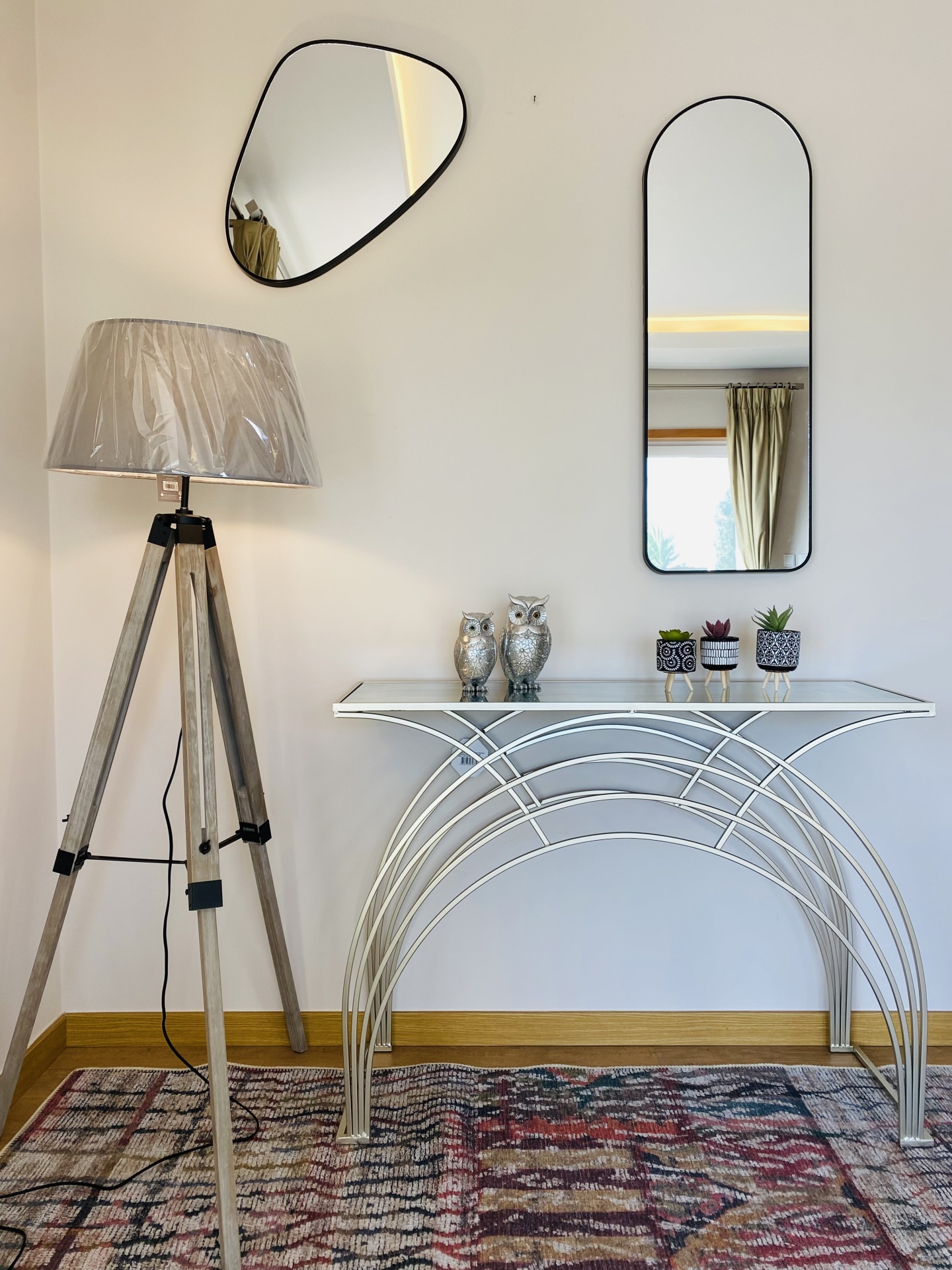 Sala com lâmpada de tripé, consola branca em metal, corujas decorativas, pequenos vasos com plantas, dois espelhos na parede e tapete colorido