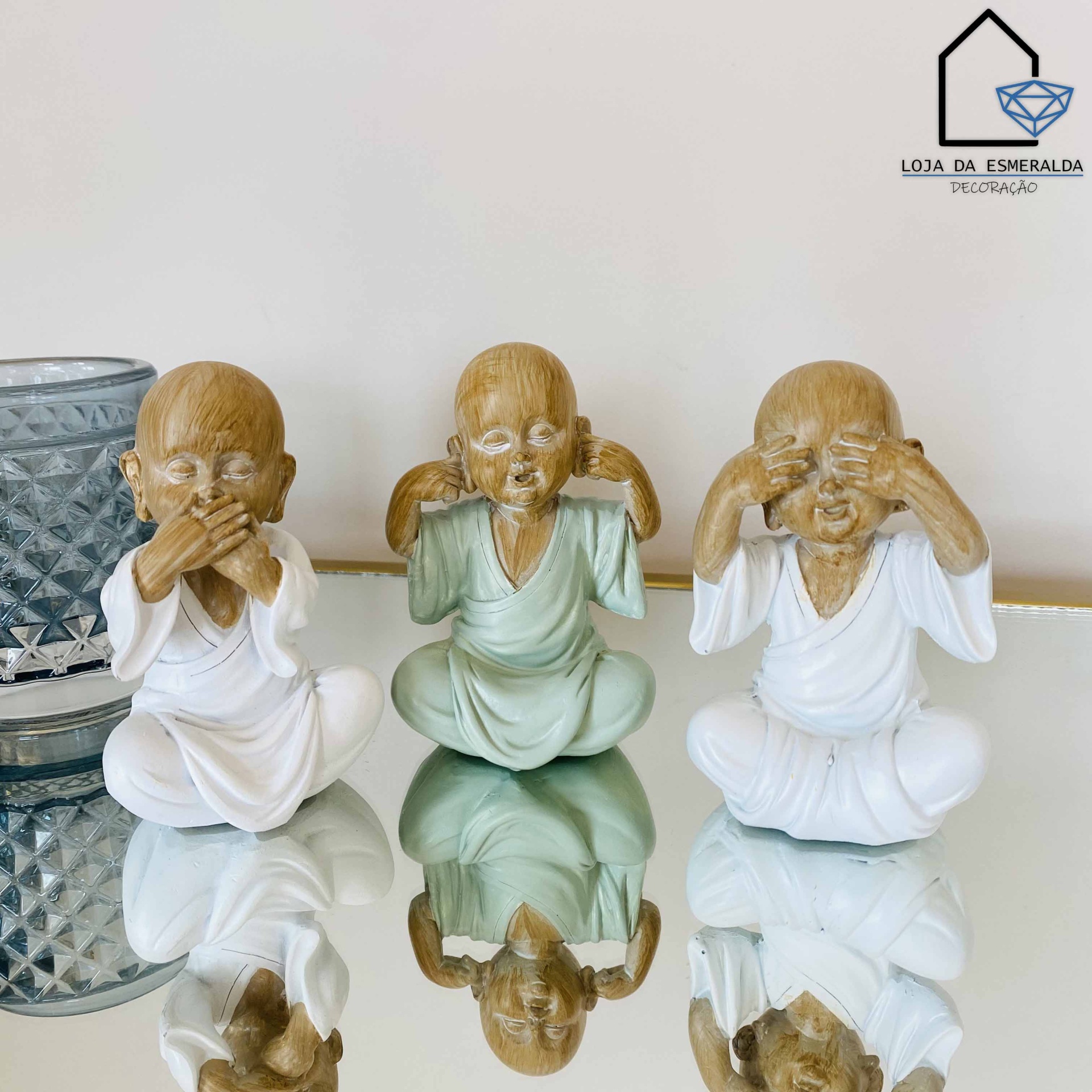 Três estatuetas de crianças em posição meditativa com gestos de não ouvir, não falar e não ver sobre mesa espelhada.