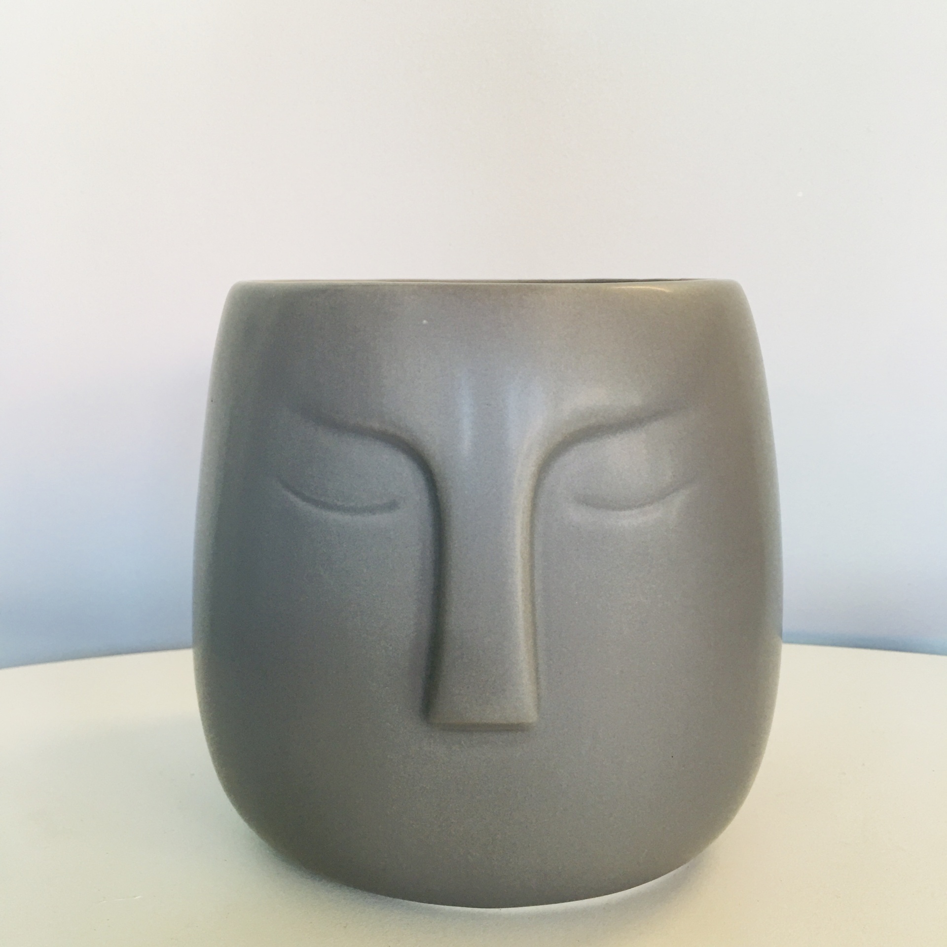 Vaso cinzento fosco com rosto estilizado em relevo sentado numa superfície branca
