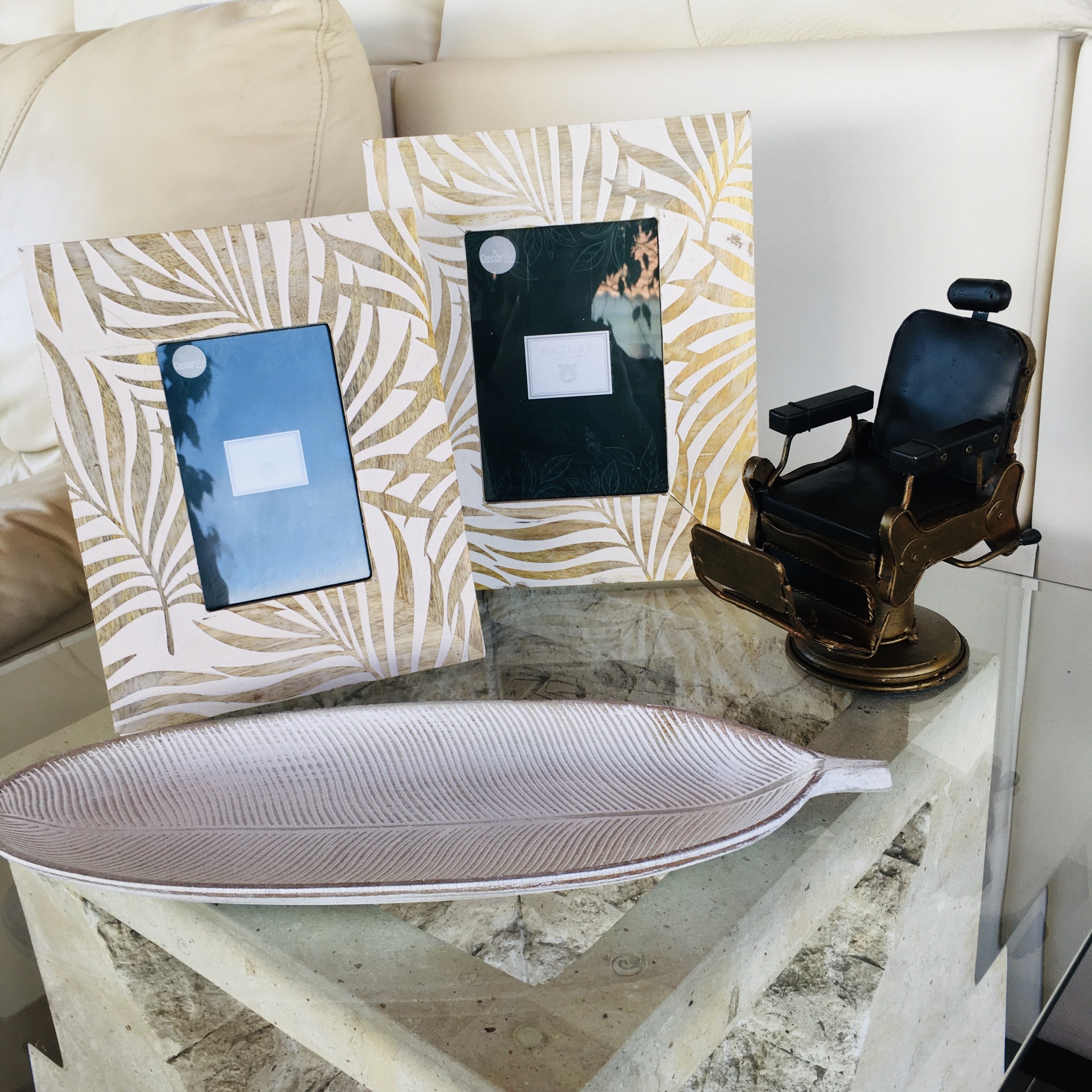 Duas molduras bege com padrão dourado, bandeja decorativa clara em forma de folha e miniatura de cadeira de barbeiro preta sobre mesa de vidro