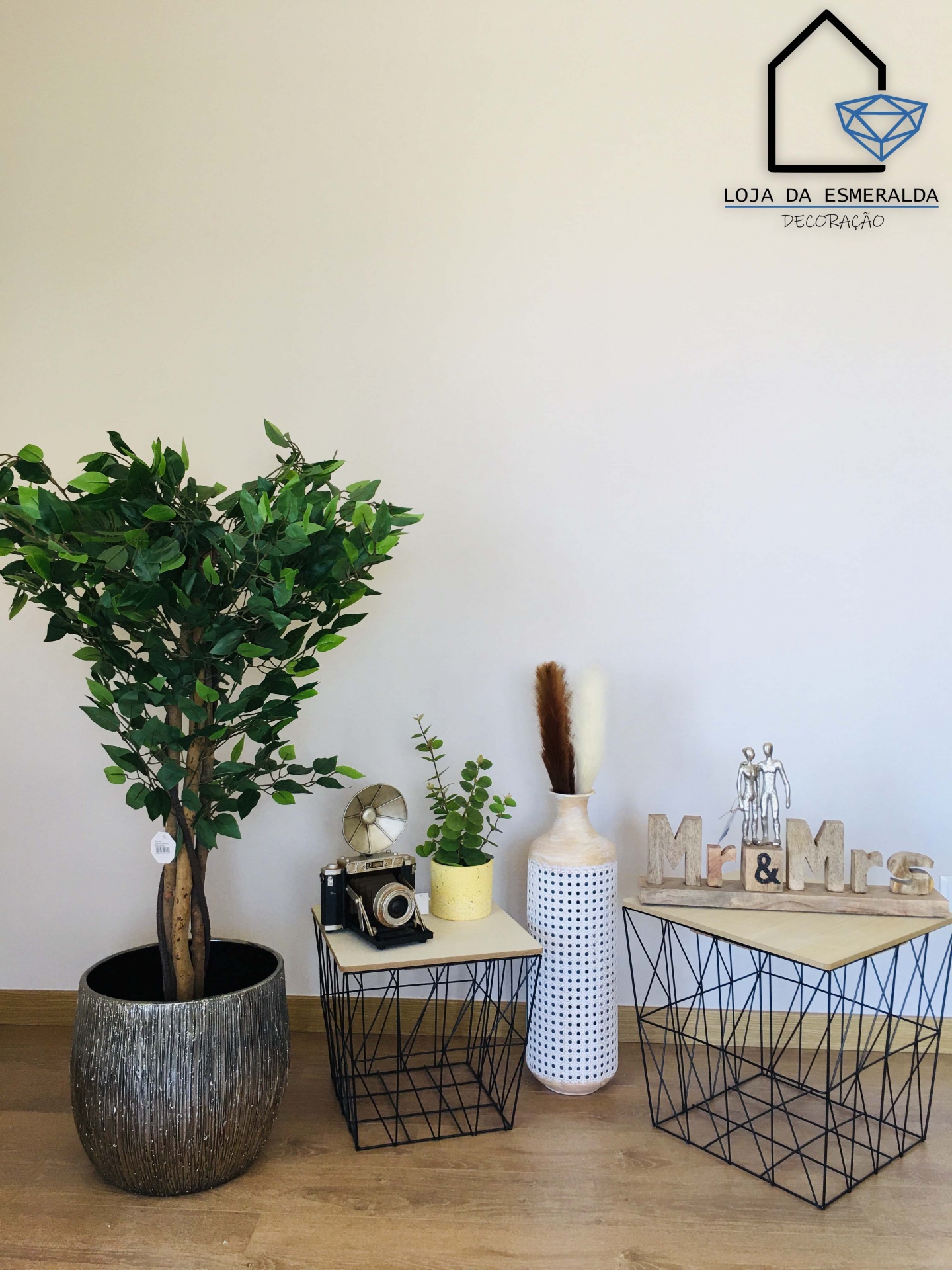 Planta artificial em vaso cinzento ao lado de mesas de apoio com objetos decorativos, jarro branco perfurado e máquina fotográfica retro