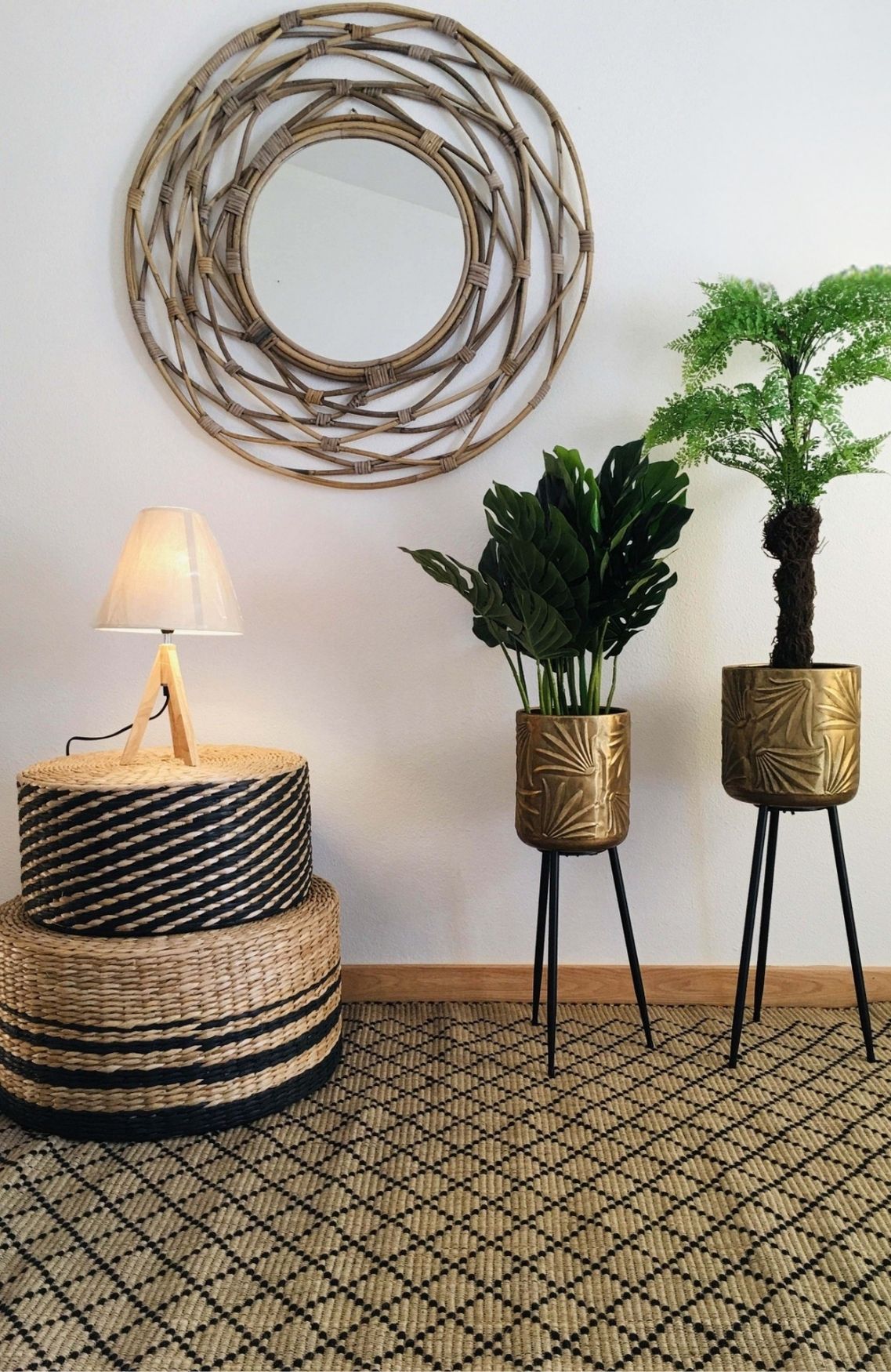 Conjunto decorativo interior com mesa de apoio em vime, candeeiro, espelho rattan, vasos metálicos dourados com plantas e tapete geométrico.