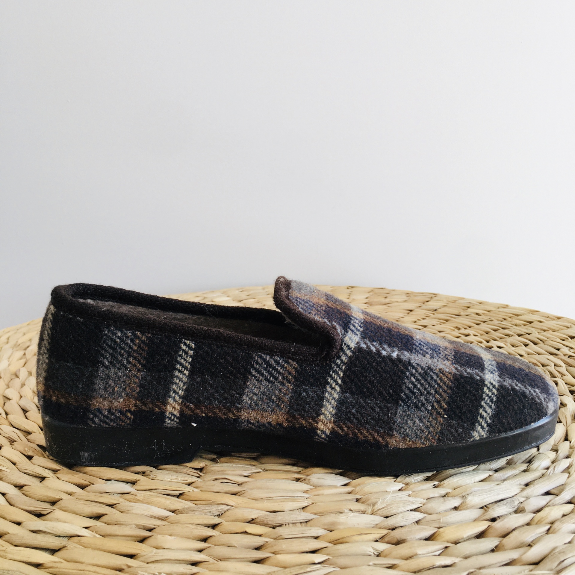 Sapato tipo pantufa xadrez preto, cinza e castanho com sola preta sobre superfície de fibras naturais