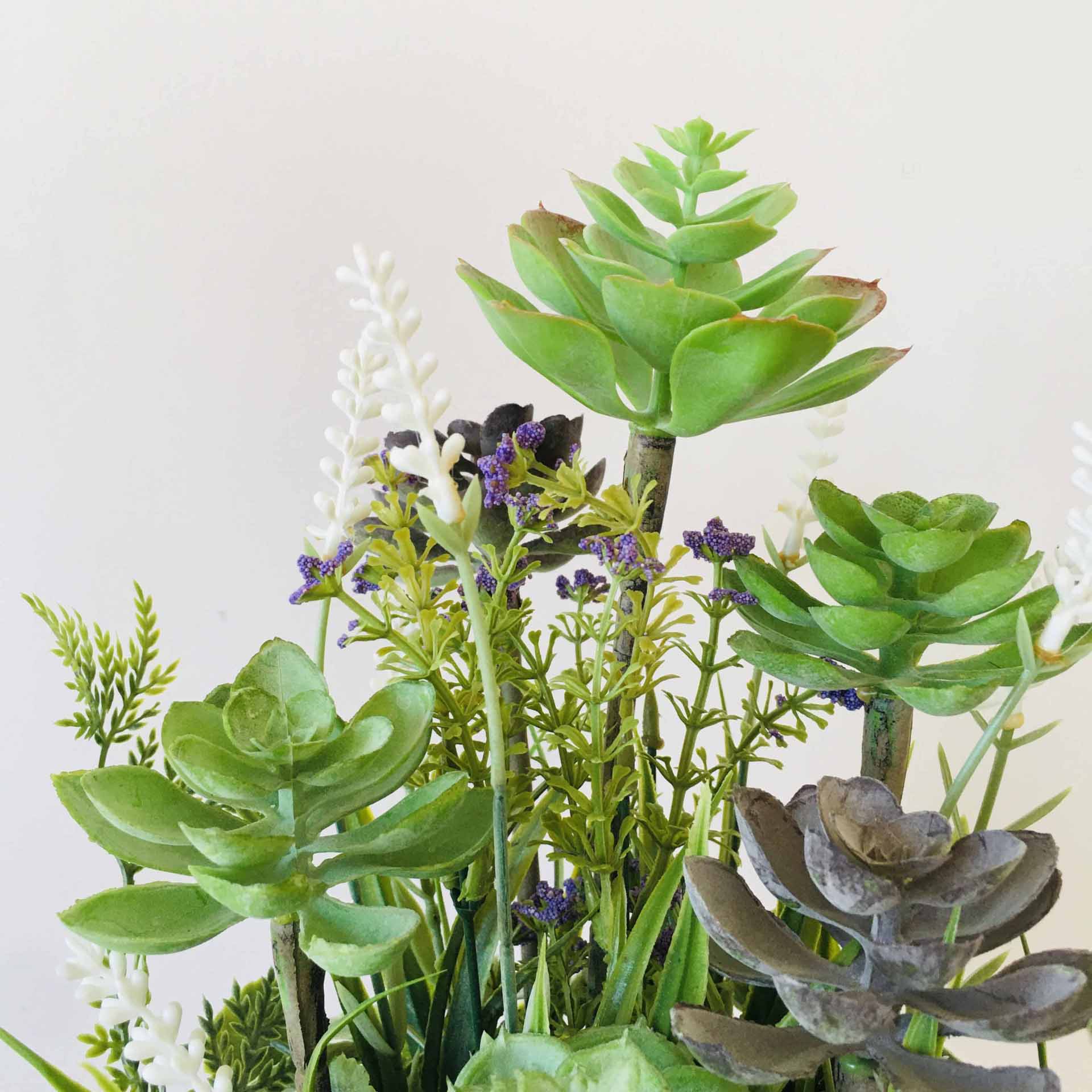 Plantas suculentas artificiais verdes e flores roxas/brancas num vaso contra fundo branco