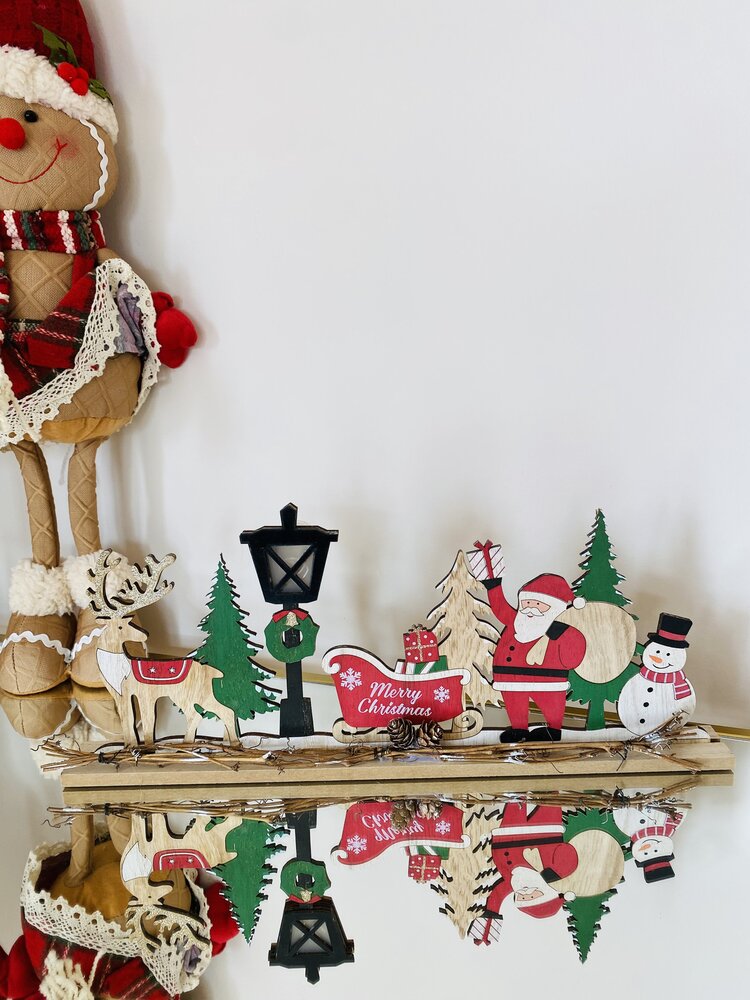 Decoração natalícia em madeira com Pai Natal, trenó, rena, árvore e boneco de neve sobre superfície reflexiva