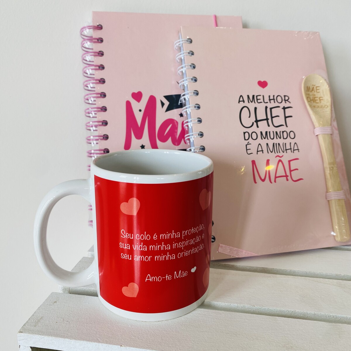 Caneca vermelha com texto, dois cadernos rosa, colher de madeira e elástico rosa sobre superfície branca.