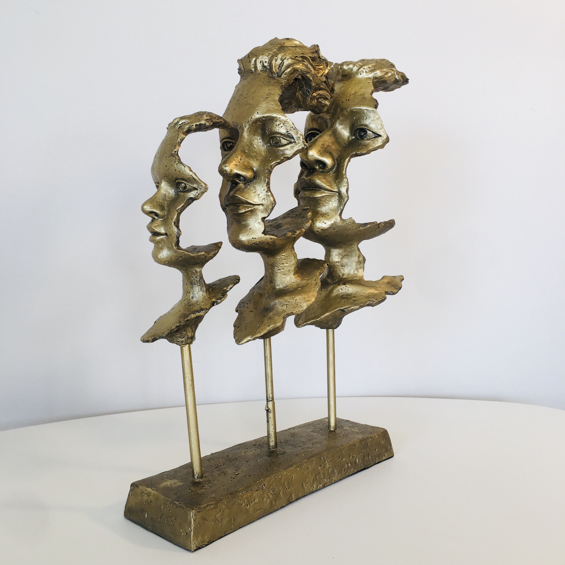 Escultura dourada com três rostos estilizados em metal sobre base retangular