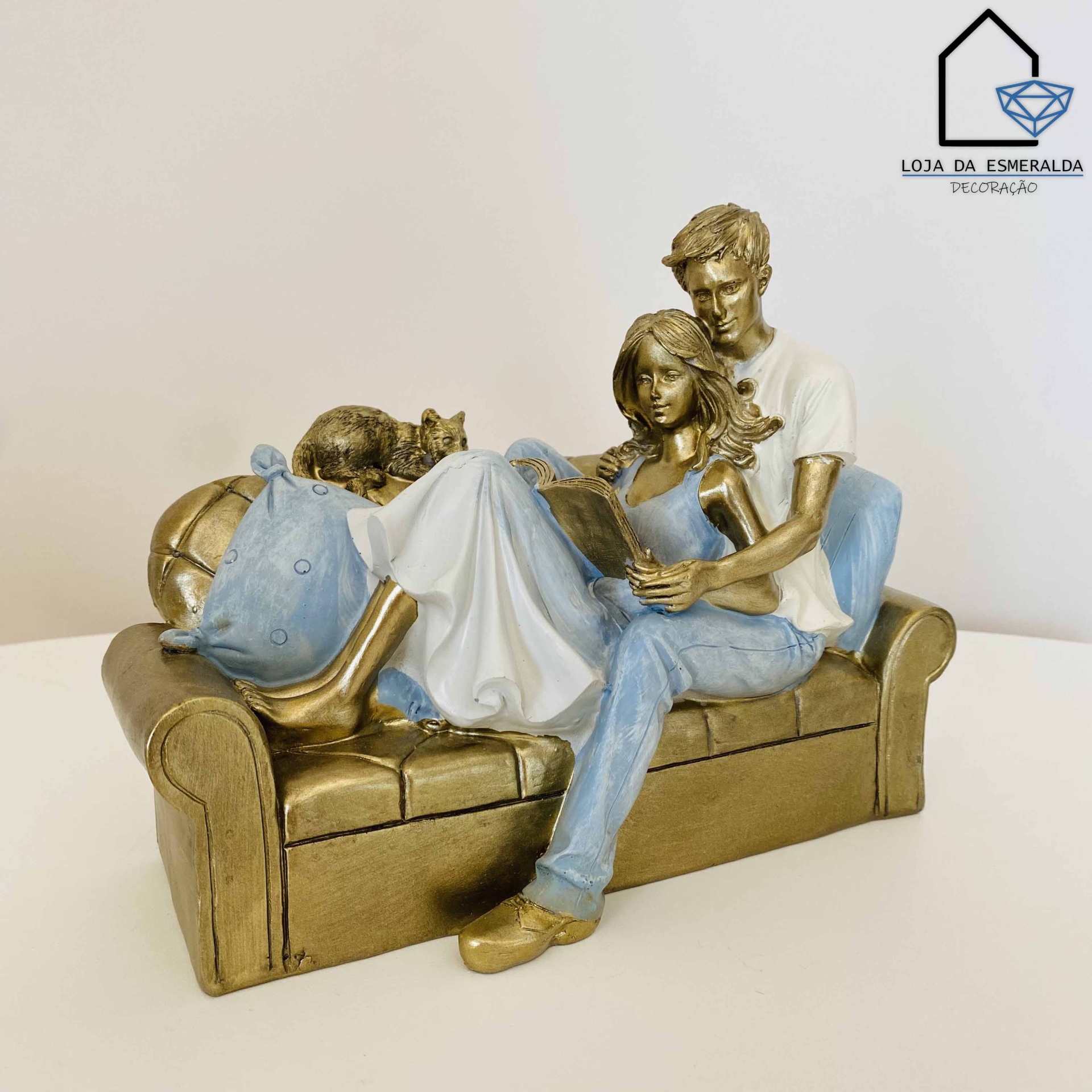Escultura de casal sentado num sofá com gato e almofada, cores castanho dourado, azul e branco.