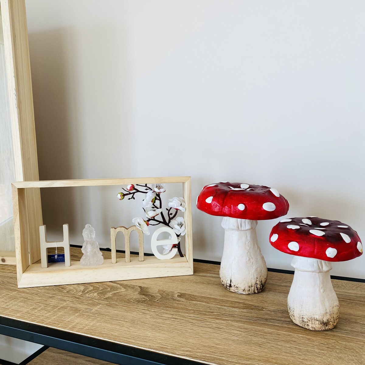 Decoração sala com moldura de madeira e letras m e, estátua e ramo, dois cogumelos vermelhos com pintas brancas sobre mesa de madeira.