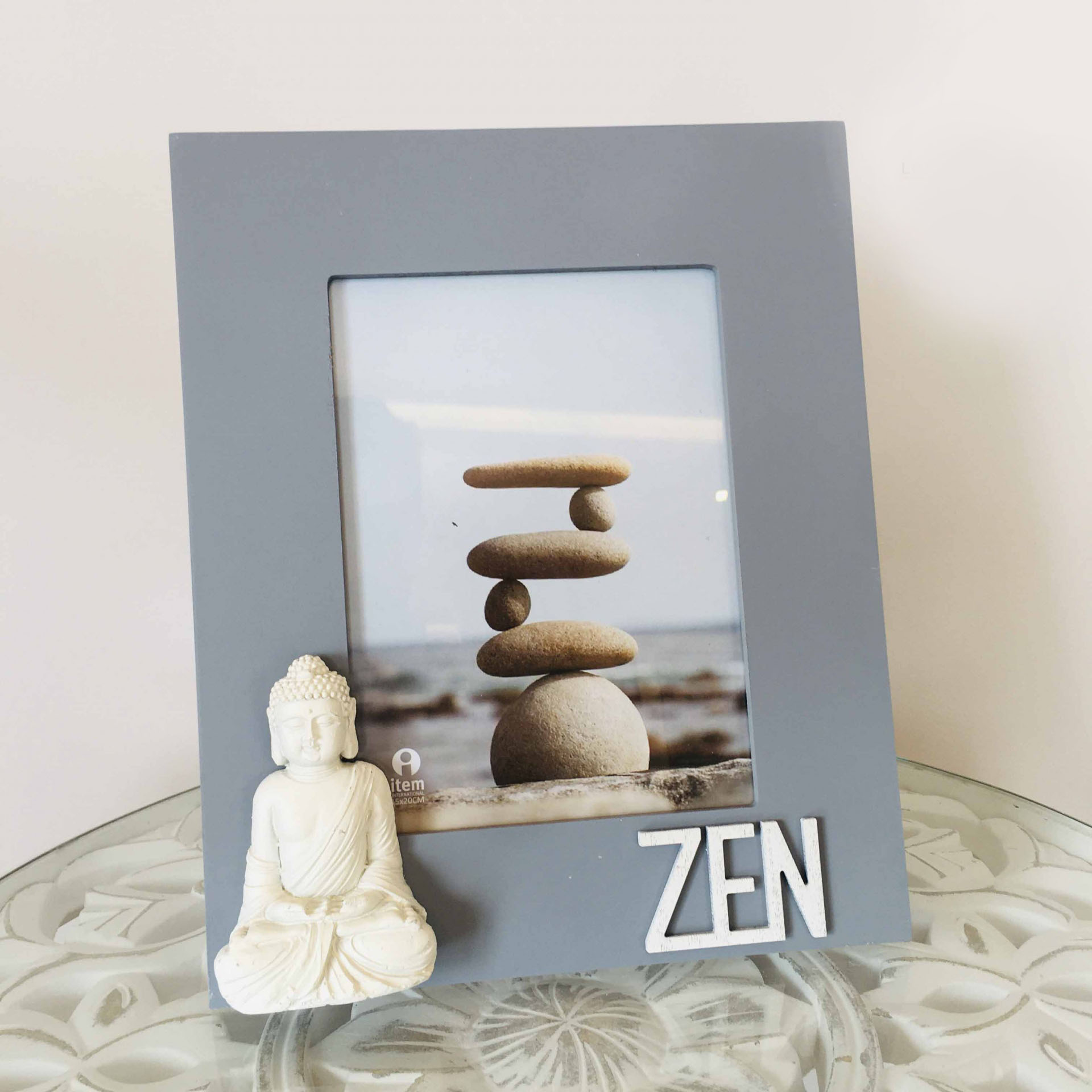 Moldura cinza com palavra 'ZEN' e estátua branca de Buda, sobre mesa de vidro com padrão floral