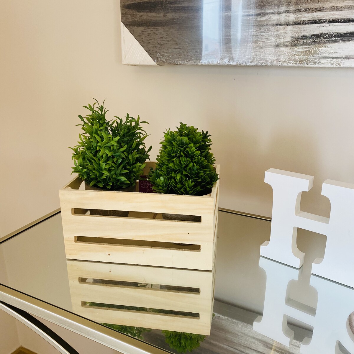 Caixa de madeira branca com plantas artificiais verdes numa mesa espelhada ao lado de letras decorativas brancas.