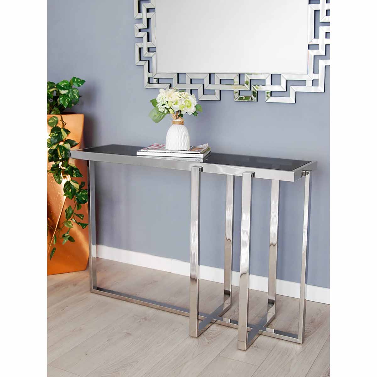 Consola moderna com tampo cinzento e estrutura metálica cromada, decorada com flores e espelho geométrico na parede.