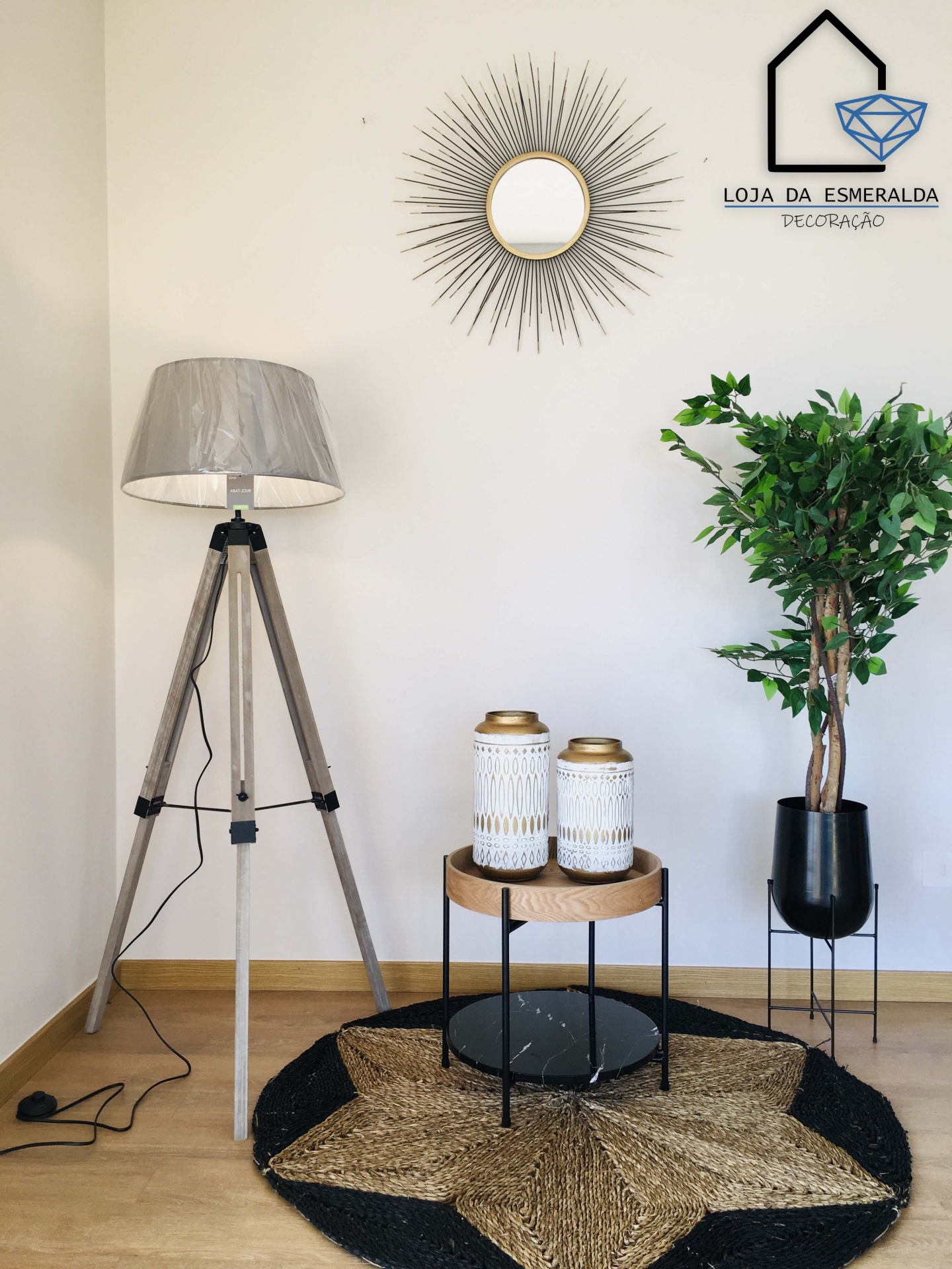 Candeeiro de chão tripé madeira com para-sol cinzento, duas lanternas brancas e douradas em mesa redonda madeira metal, tapete redondo preto castanho, planta verde vaso preto, espelho redondo moldura raios dourados