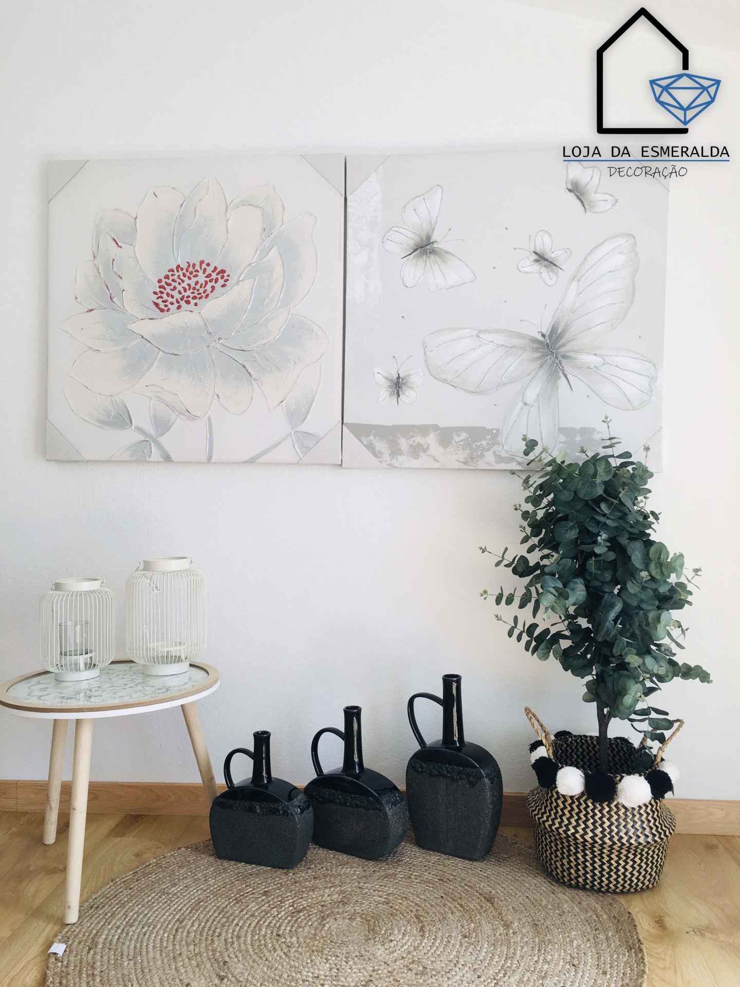 Decoração com quadros de flores e borboletas, mesa branca com candeeiros, regadores pretos, planta em vaso tecido e tapete de palha.